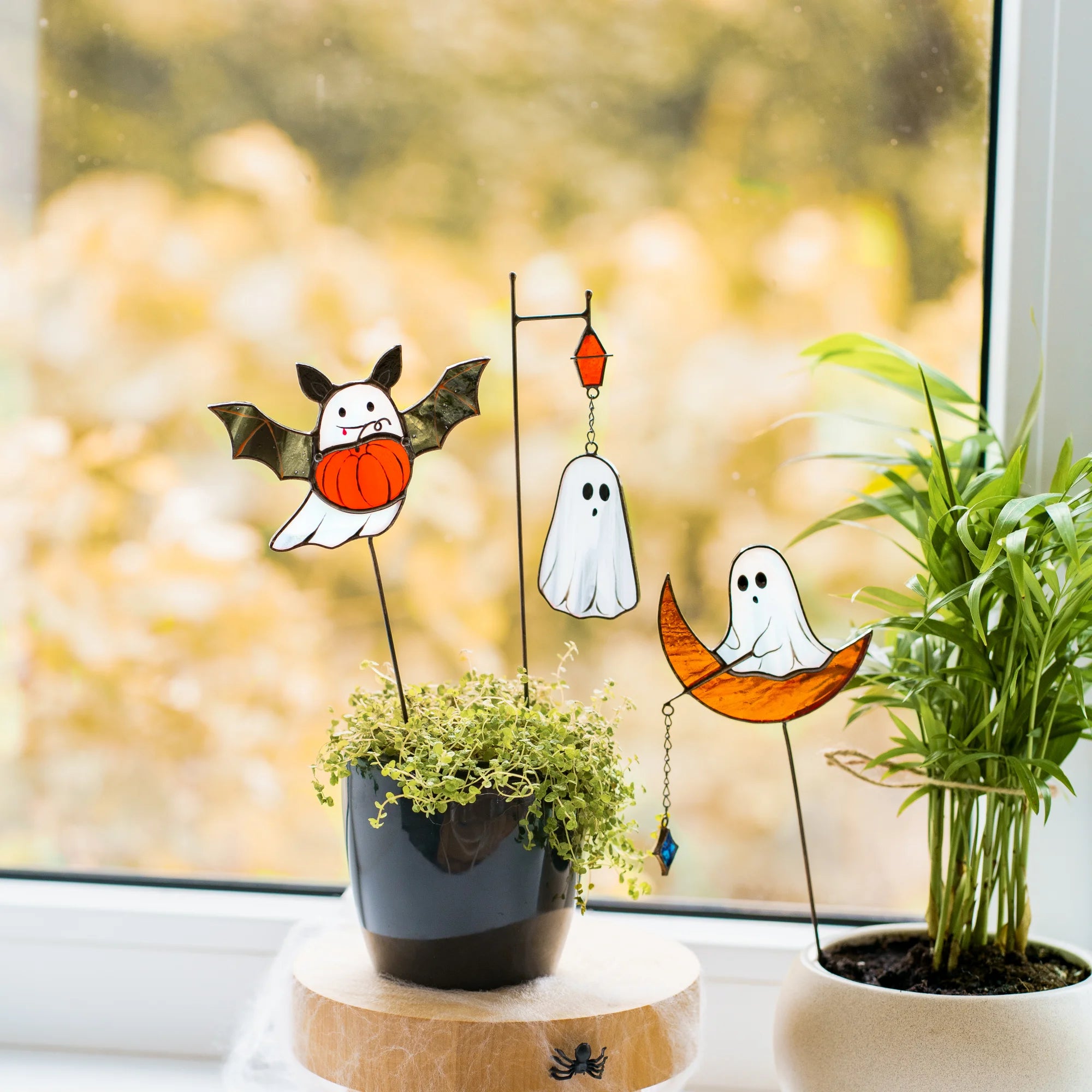 A spooky stained glass Halloween plant stake set featuring a ghost with a pumpkin and bat wings, a ghost hanging from a lantern, and a ghost sitting on a moon, perfect as a unique holiday gift.