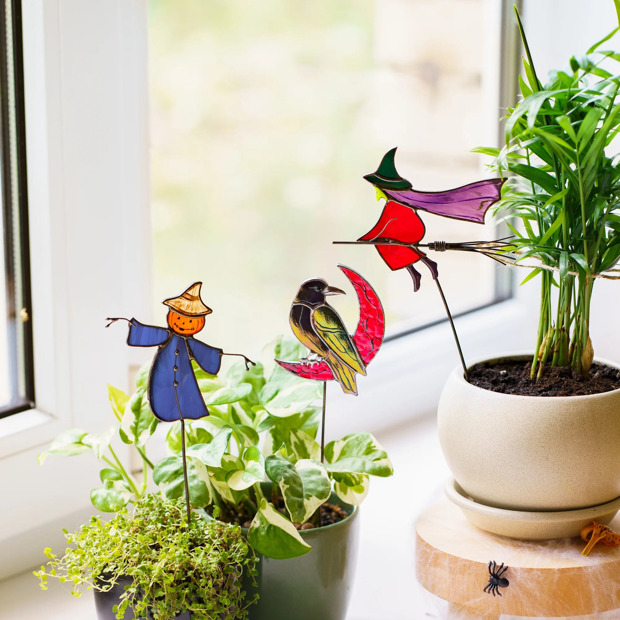 Handmade stained glass witch, raven and scarecrow plant stakes tucked into a pothos and a palm on a bright windowsill - spooky garden art for indoor plants and patio pots