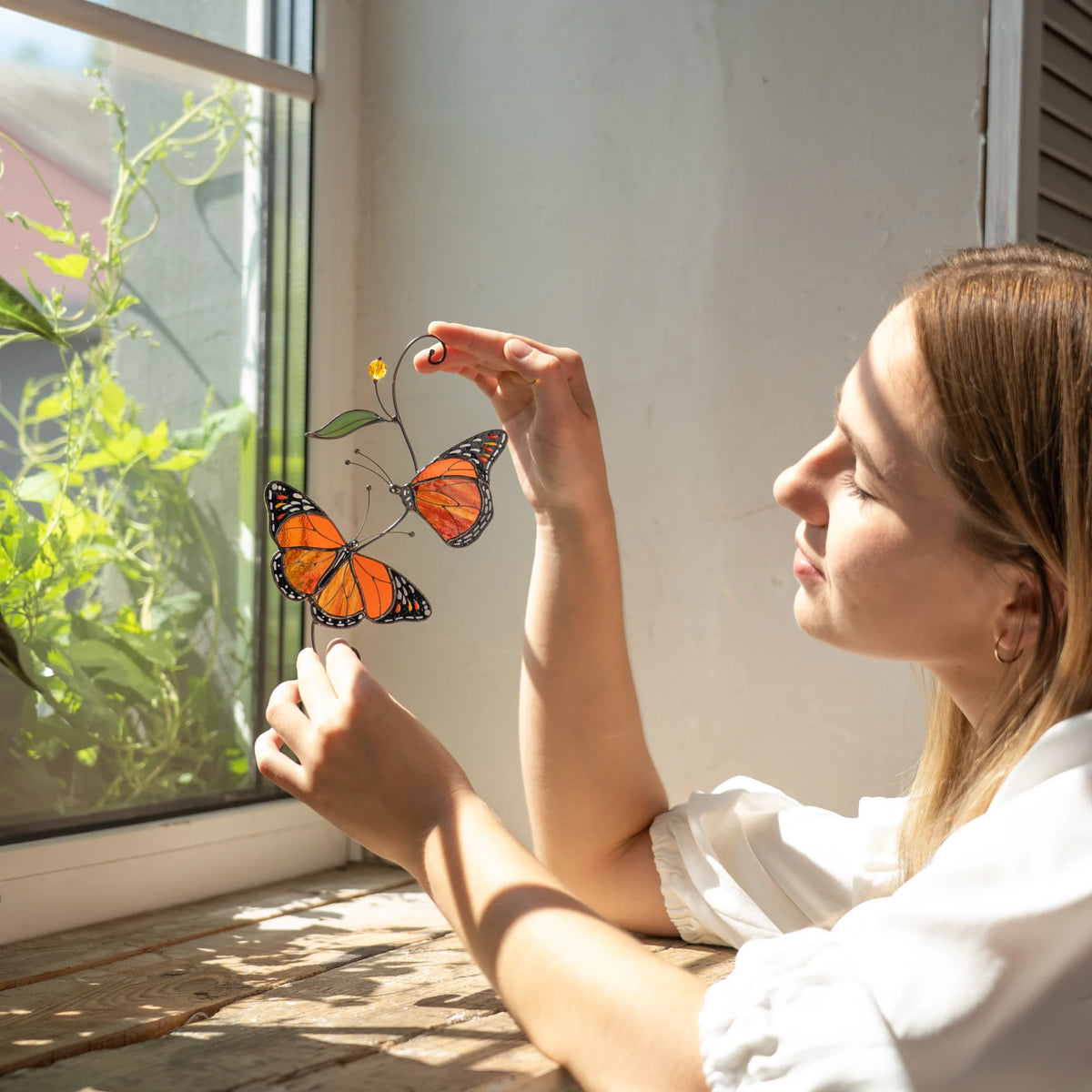 Woman at a sunlit window holding a handmade monarch butterfly stained glass suncatcher, green vines beyond the glass and warm light through the orange cathedral wings