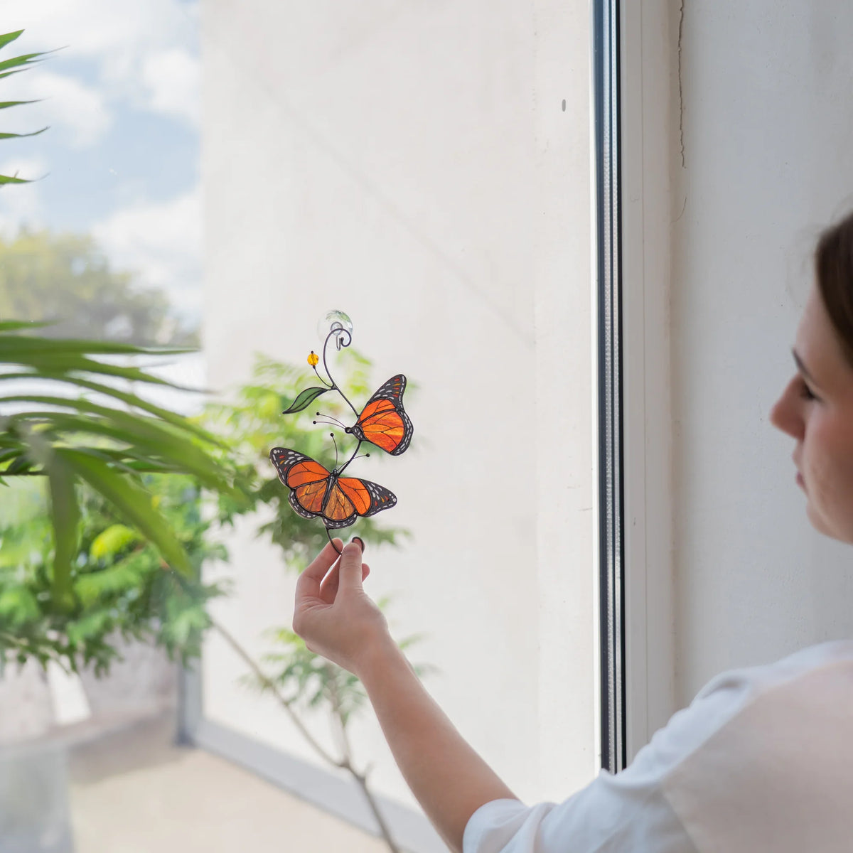 Woman hanging a monarch butterfly stained glass suncatcher on a bright window with a clear suction cup, a green palm in front - handmade decor and a gift for nature lovers