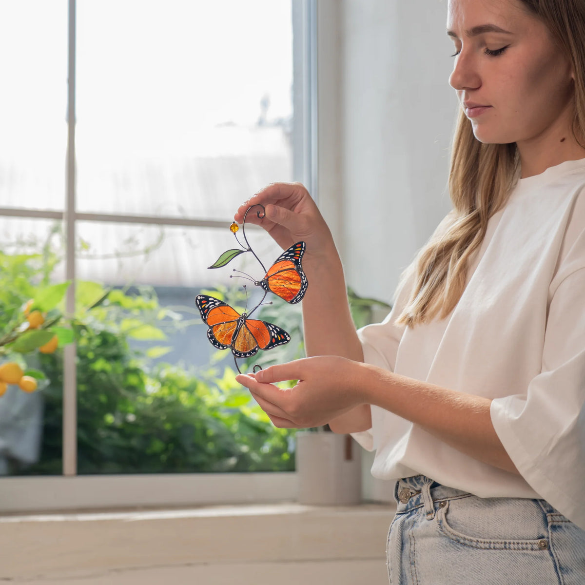 Woman studying a handmade monarch butterfly suncatcher by a window with a lemon tree behind her - stained glass wings in orange and amber edged with fine white dots