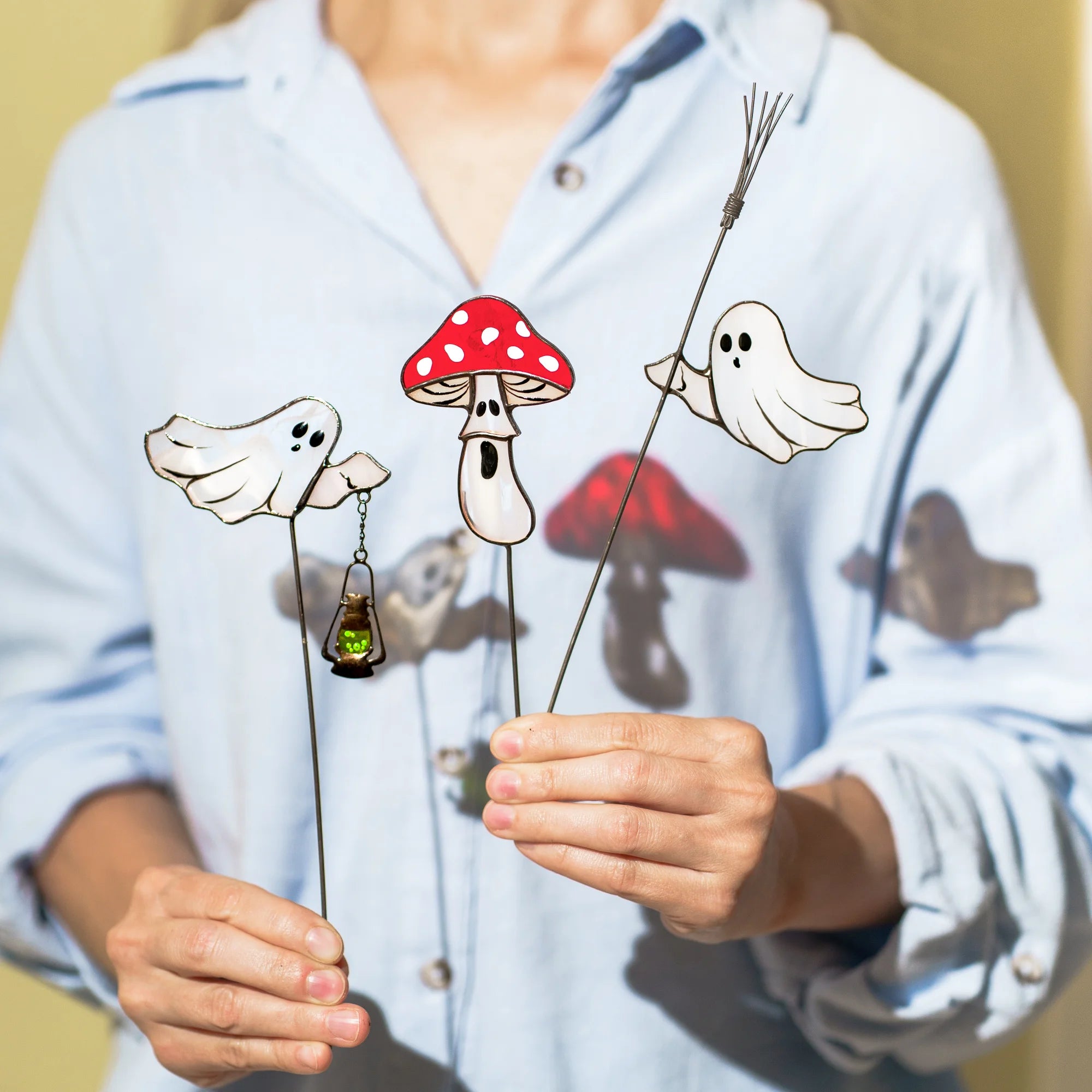 Woman holding all three handmade stained glass plant stakes - ghost with a green lantern, red toadstool mushroom and ghost on a twig broom, showing their slim dark metal stakes