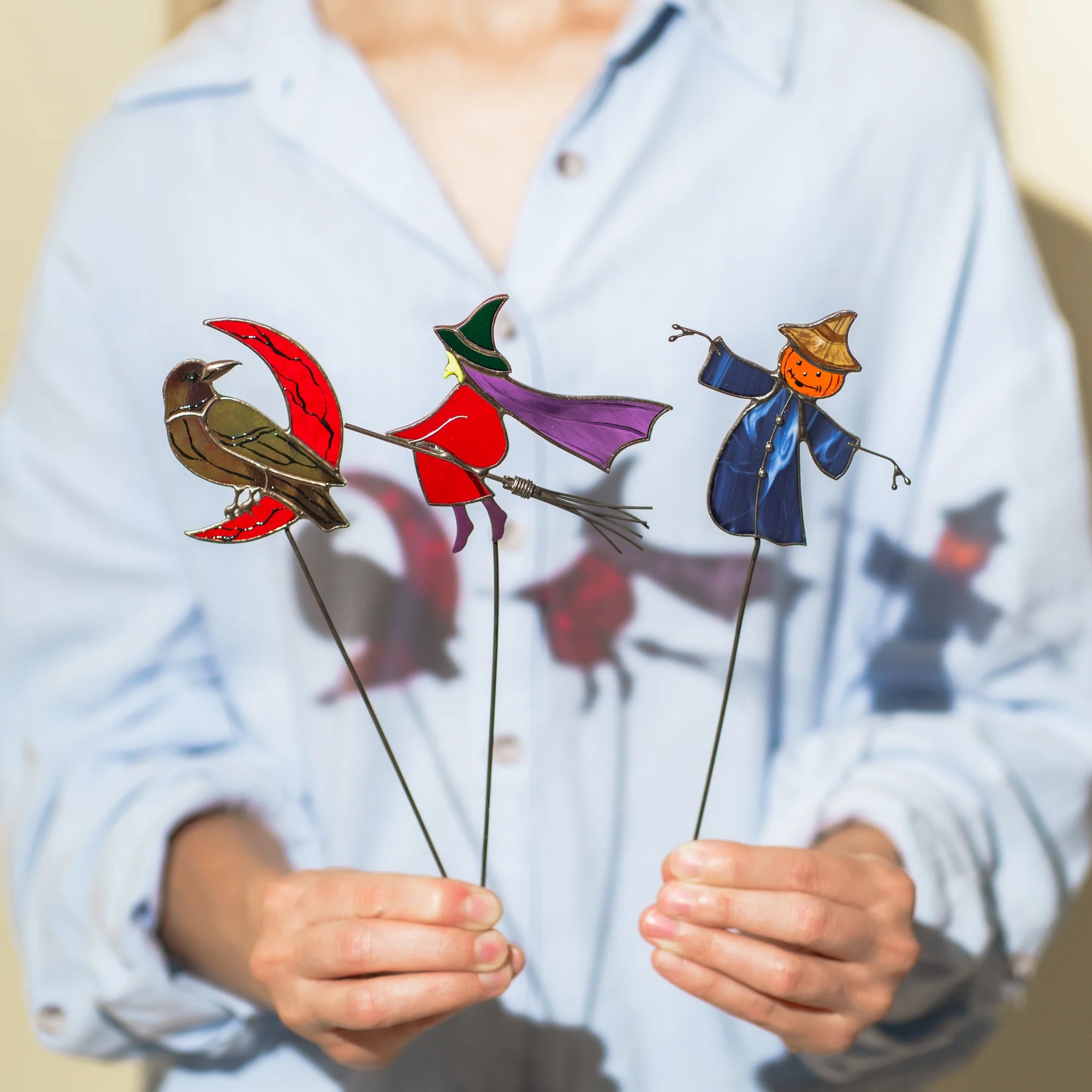 Woman holding three handmade stained glass plant stakes - a raven on a red crescent moon, a witch in a violet cape on a wire broom and a pumpkin-headed scarecrow in blue