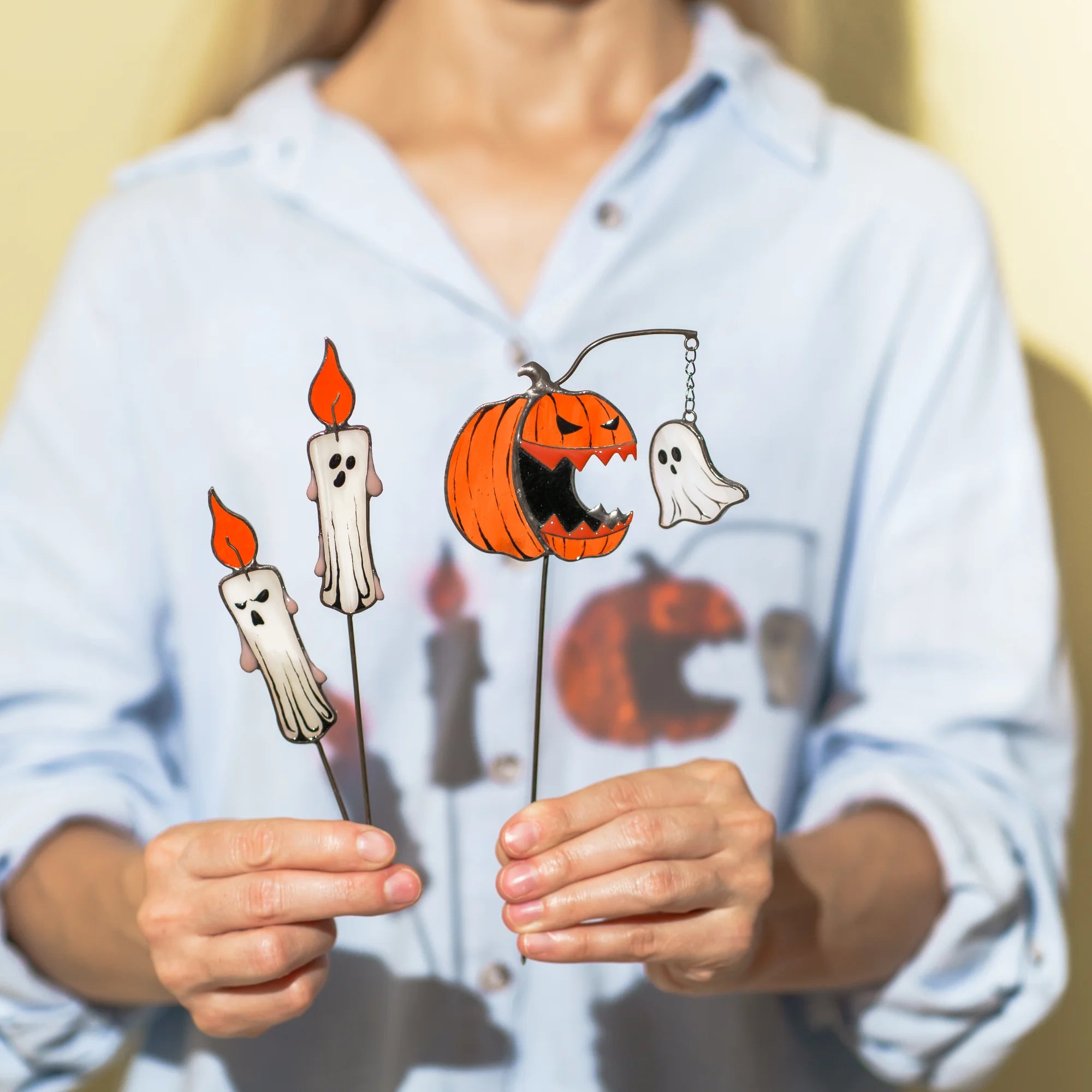 A person wearing a light blue button-up shirt holds three Halloween-themed stained galss plant stakes: two candle-shaped ghosts with orange flames, and one jack-o'-lantern with a hanging ghost.