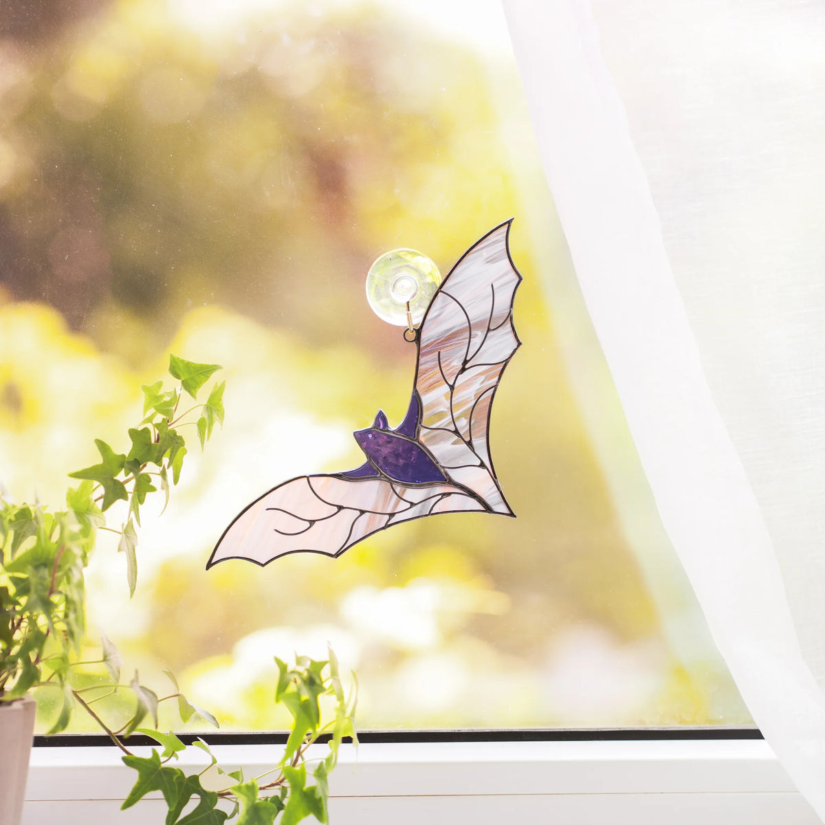 Purple bat stained glass suncatcher hanging on a suction hook in a bright window above an ivy plant - handmade Halloween window decor for a kitchen or living room