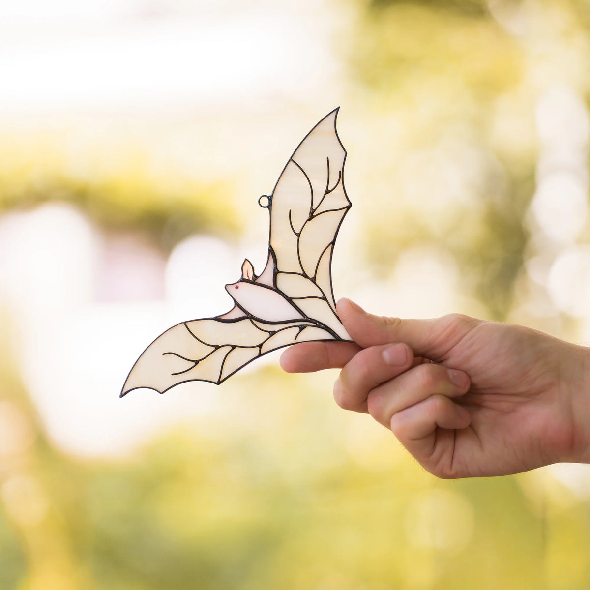 Hand holding the white stained glass bat suncatcher by its wingtip, with soft yellow-green bokeh foliage blurred behind it.