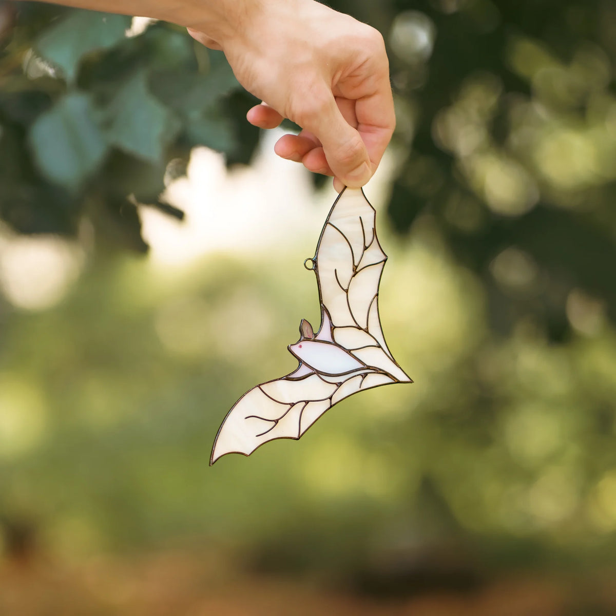White stained glass bat suncatcher dangling from a hand's fingertips in front of lush green leafy foliage on a sunny day.
