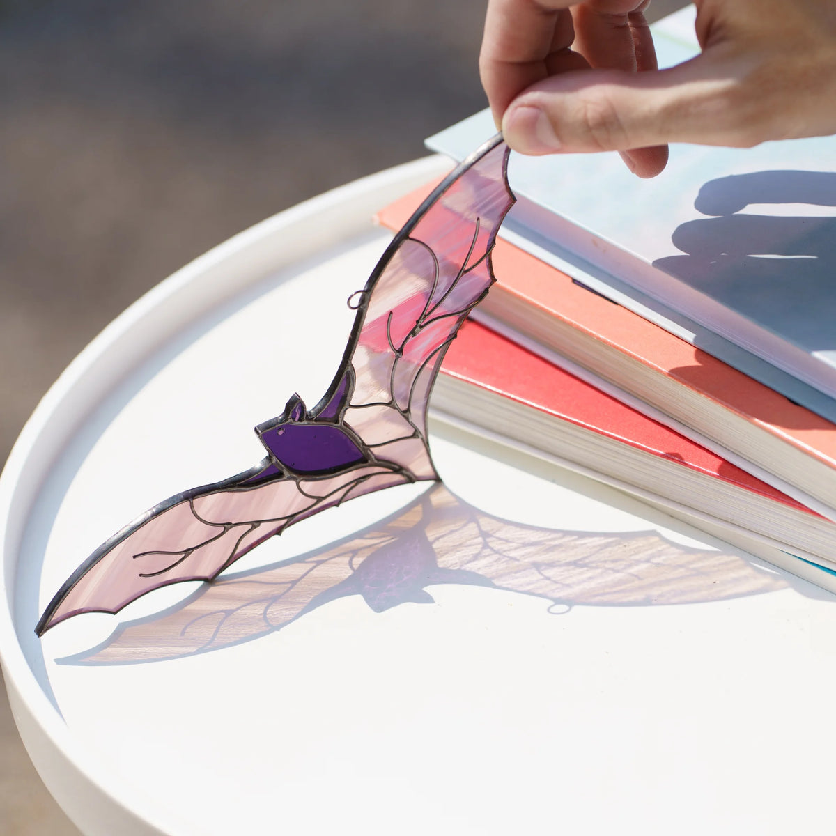 Purple bat stained glass suncatcher held above a white table with books, bright sun throwing a lilac shadow of the wings onto the surface - handmade Halloween decor