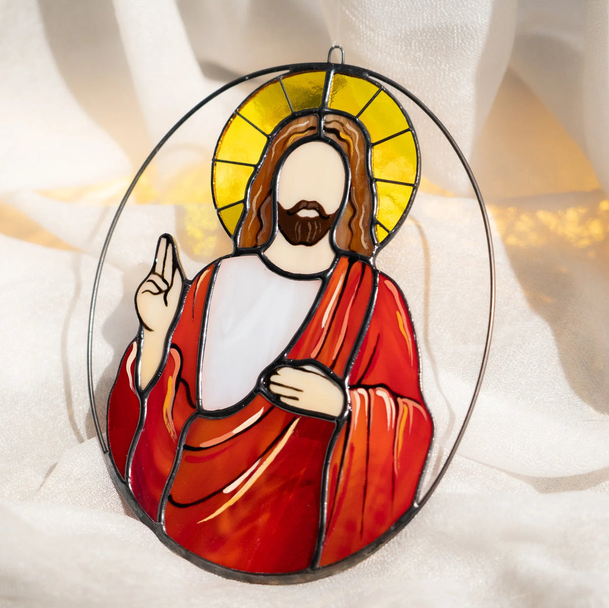 Jesus stained glass suncatcher with a golden halo, red robe and raised hand of blessing in an oval wire frame, resting on soft white fabric in warm light - handmade religious window decor