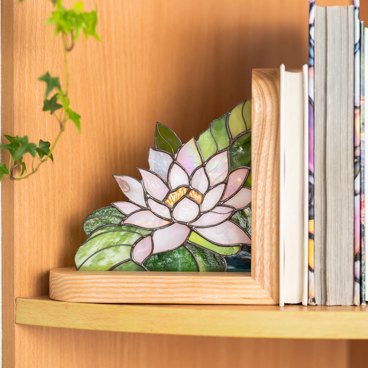 Stained glass water lily bookend resting beside a stack of books on a bookshelf, light ash wood frame glowing warm in natural daylight
