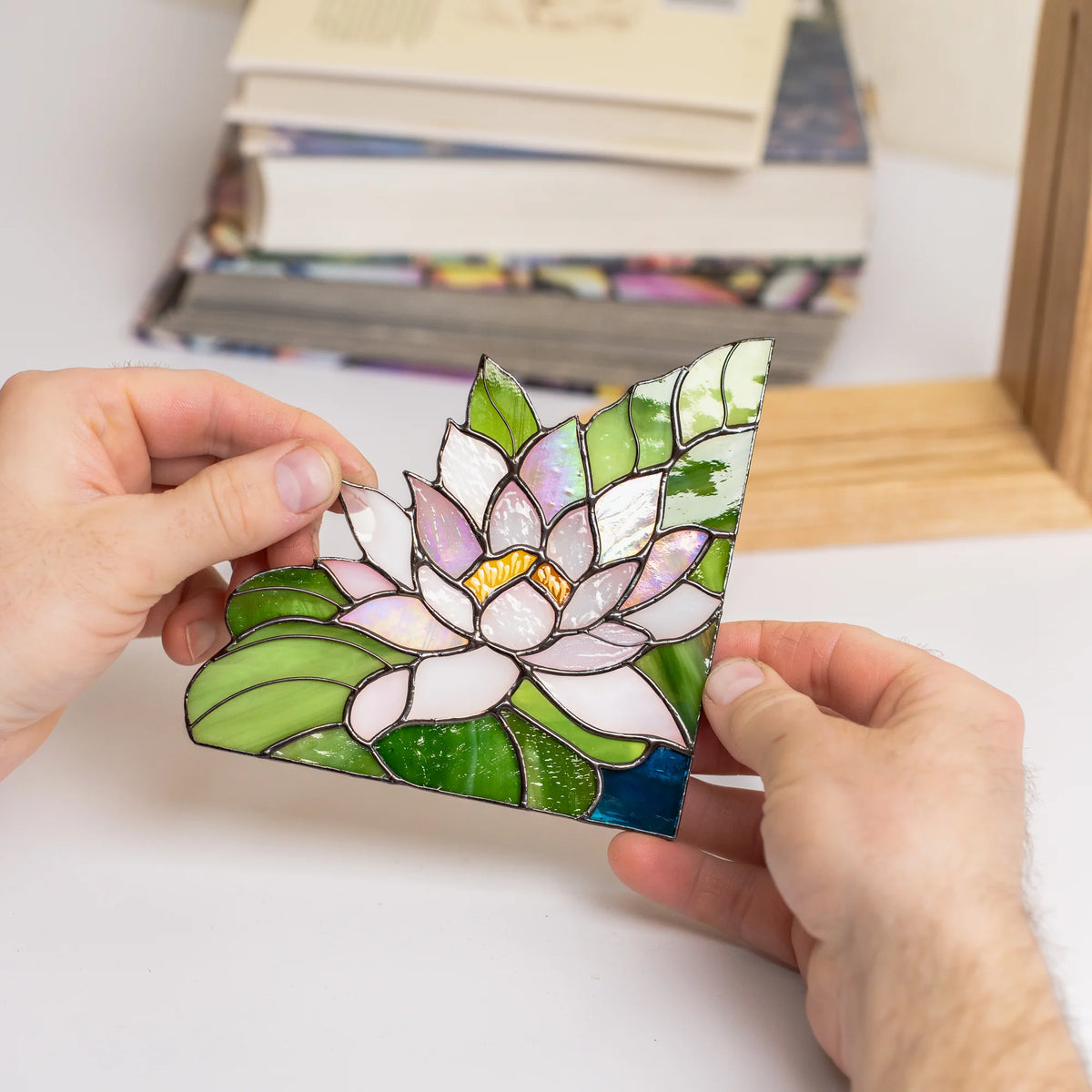 Close-up of hands placing a stained glass water lily bookend on a bookshelf, showing the soldered copper foil seams and iridescent mother-of-pearl glass petals