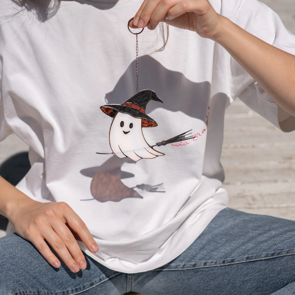 Stained glass ghost in a witch's hat held close against a white t-shirt, showing the true scale of this handmade Halloween suncatcher with broom in hand