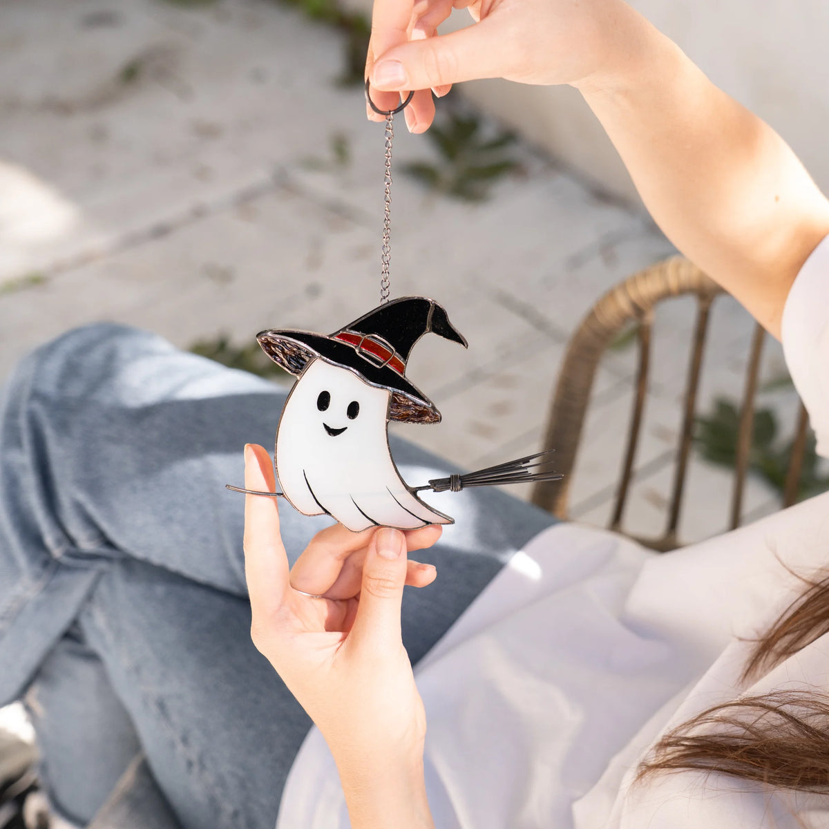 Hand holding a stained glass Halloween ghost suncatcher outdoors near a rattan chair and paving stones, a playful handmade decoration for fall and Halloween
