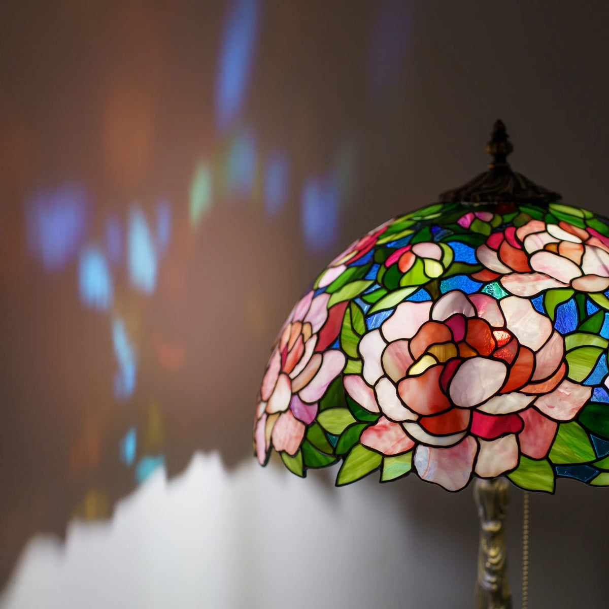 Close-up of a stained glass peony lamp shade glowing with pink and green glass petals, soft blue and amber light reflections blurred behind it, handmade Tiffany technique table lamp