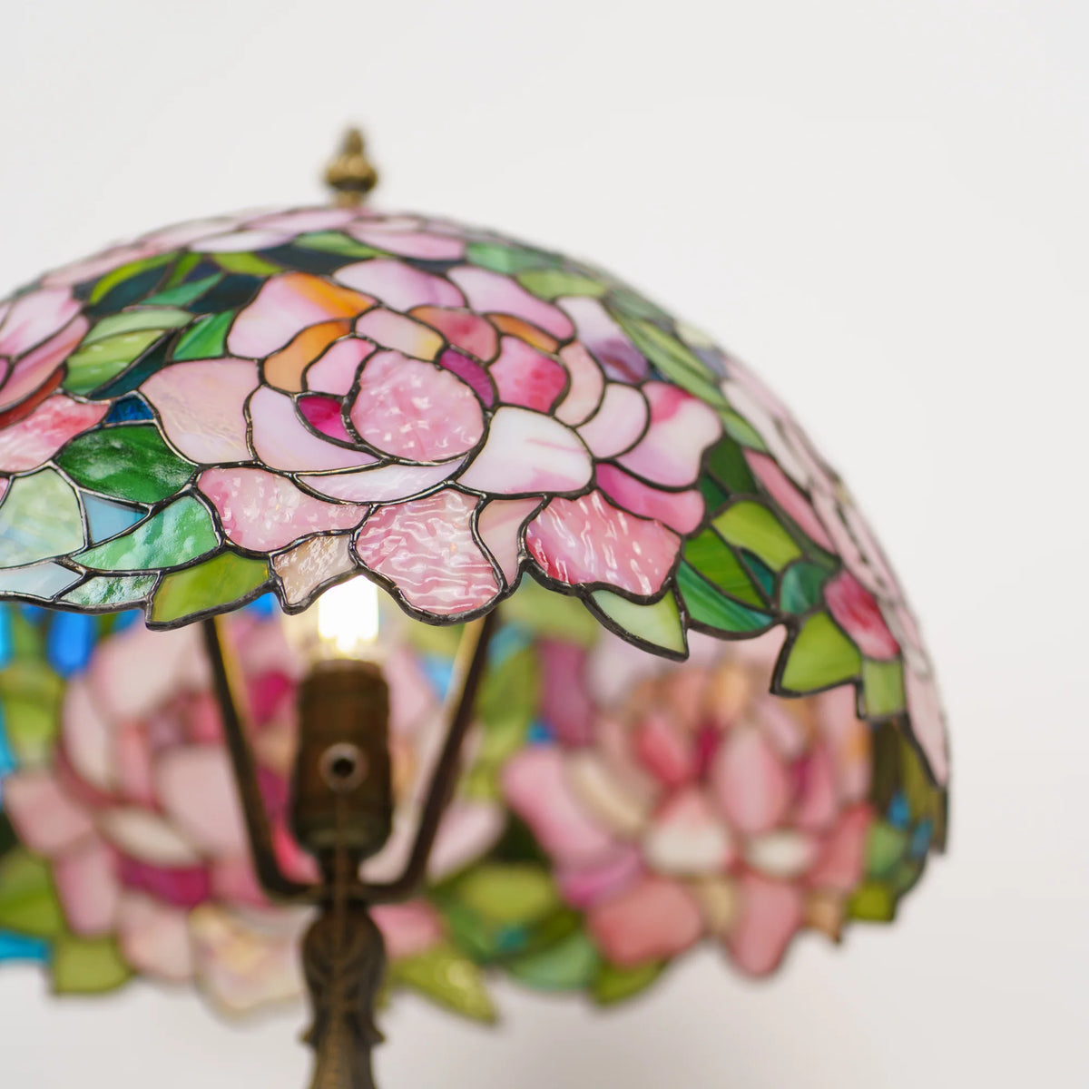 Looking up inside a stained glass peony table lamp at the glowing bulb and brass socket, pink and green glass petals and blue accents visible through the shade - handmade Tiffany style lamp