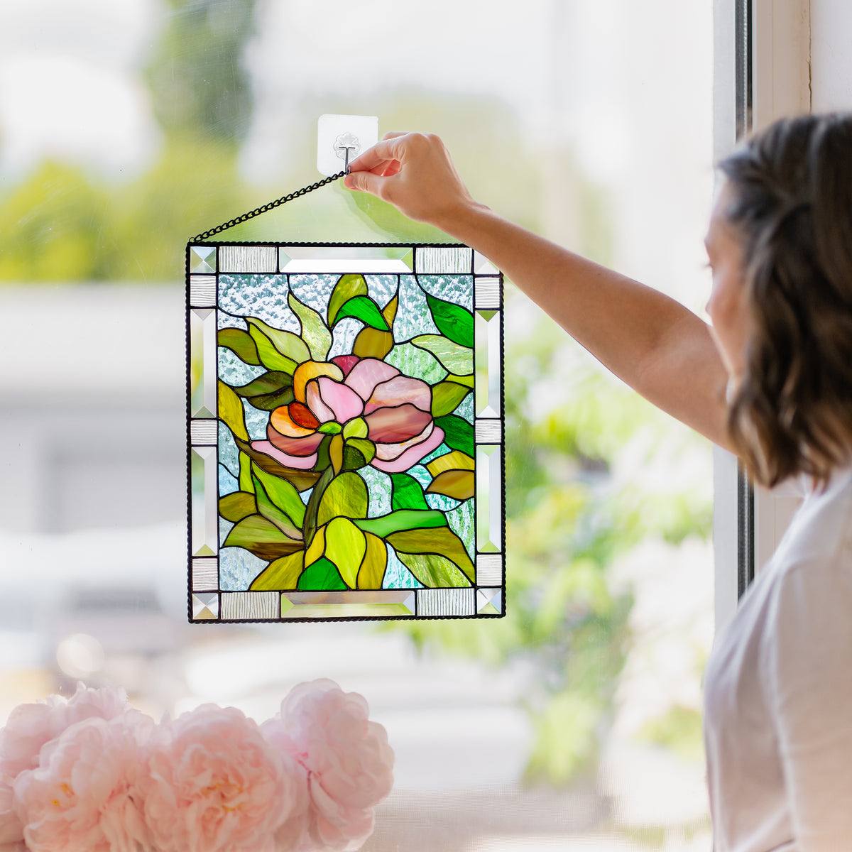 Woman holding a stained glass peony panel by its chain while sunlight casts colorful reflections on the floor beside her.
