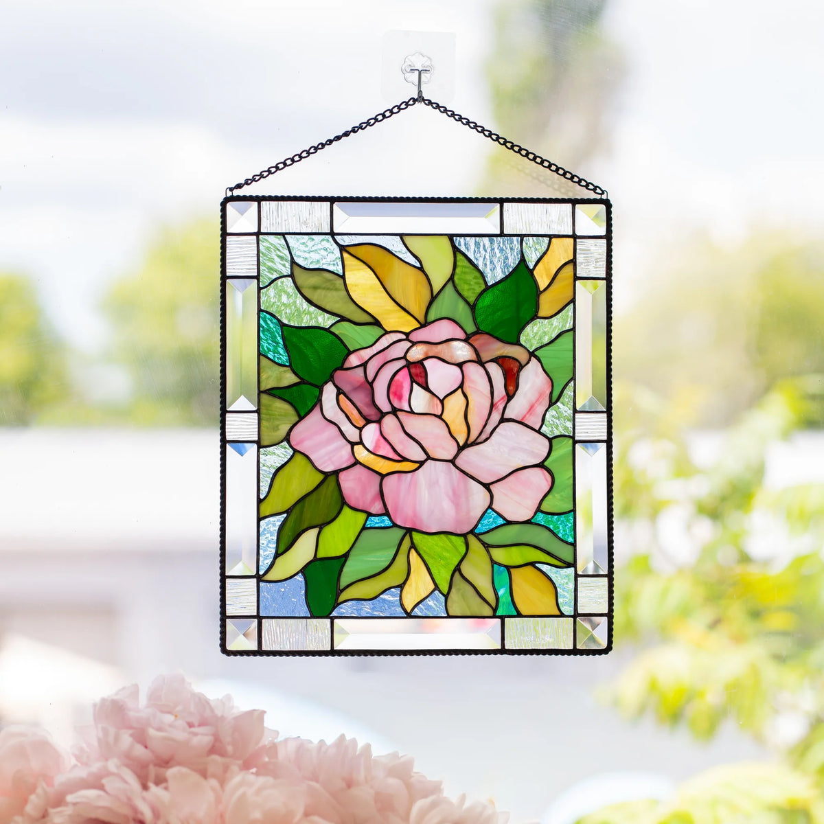 Stained glass pink peony panel with a beveled clear glass border hanging by a chain from a suction cup hook on a sunlit window.