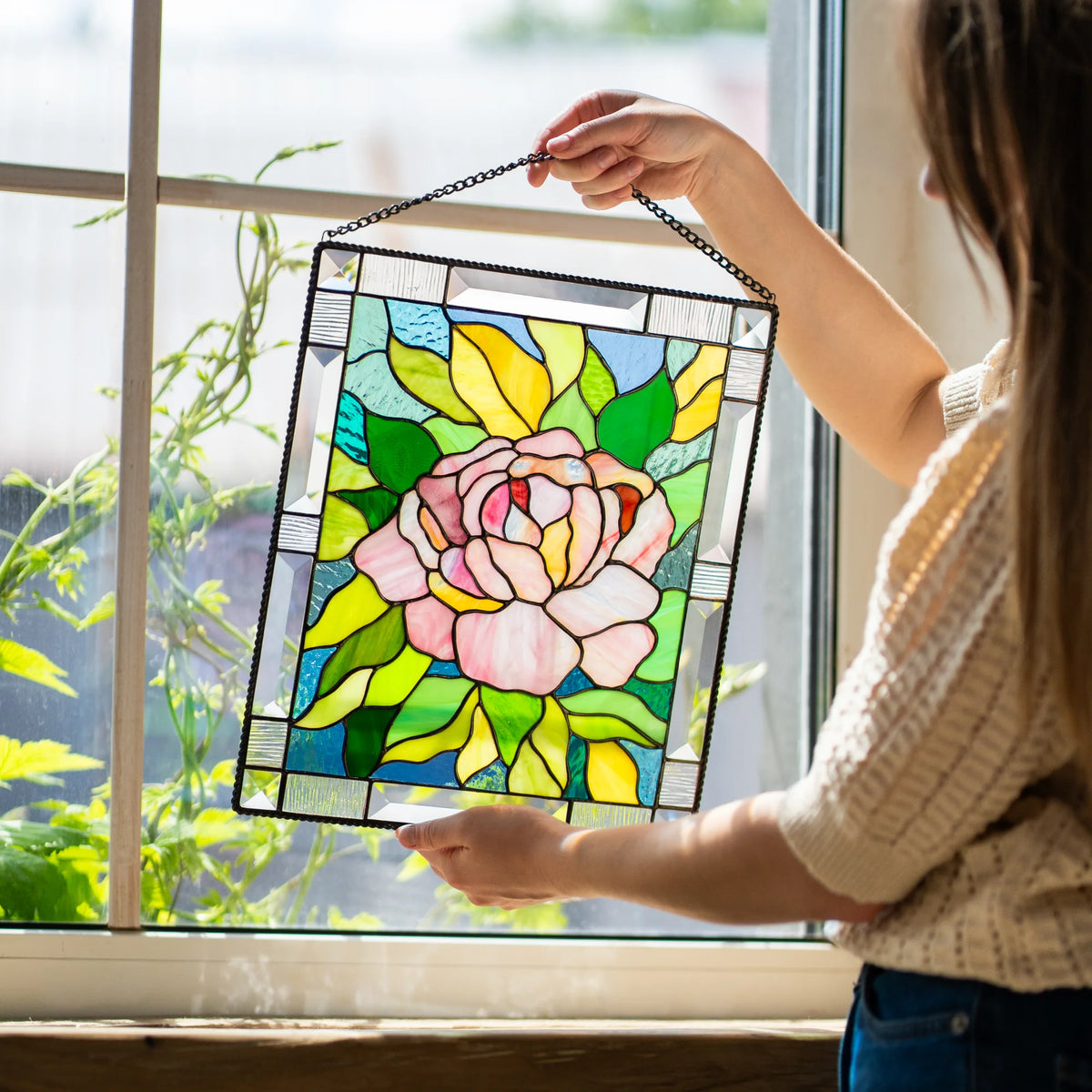 Woman holding a stained glass pink peony panel with both hands in front of a sunlit window beside a climbing green houseplant.