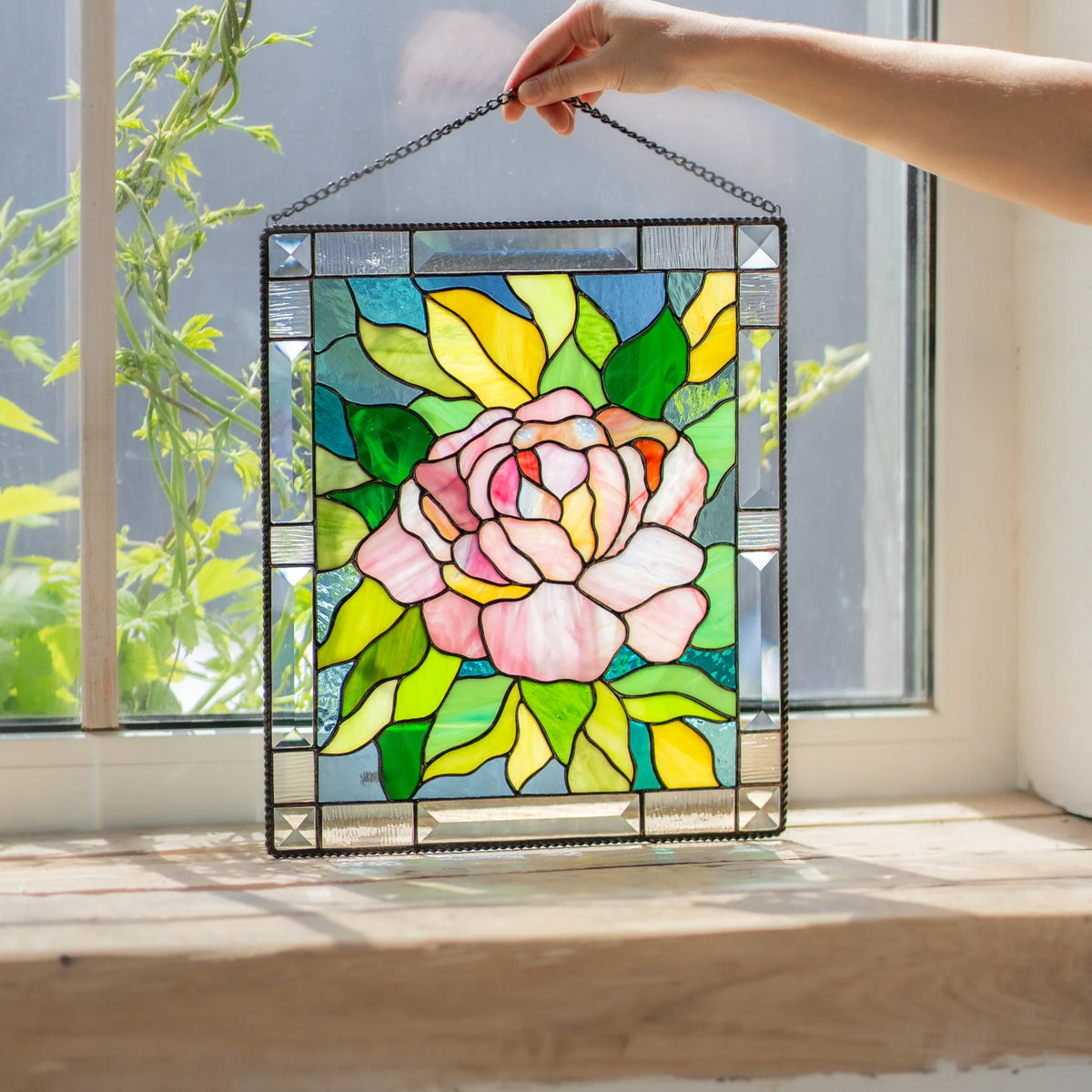 Hand holding the chain of a stained glass peony panel against a bright window, showing the beveled border and pink flower design.