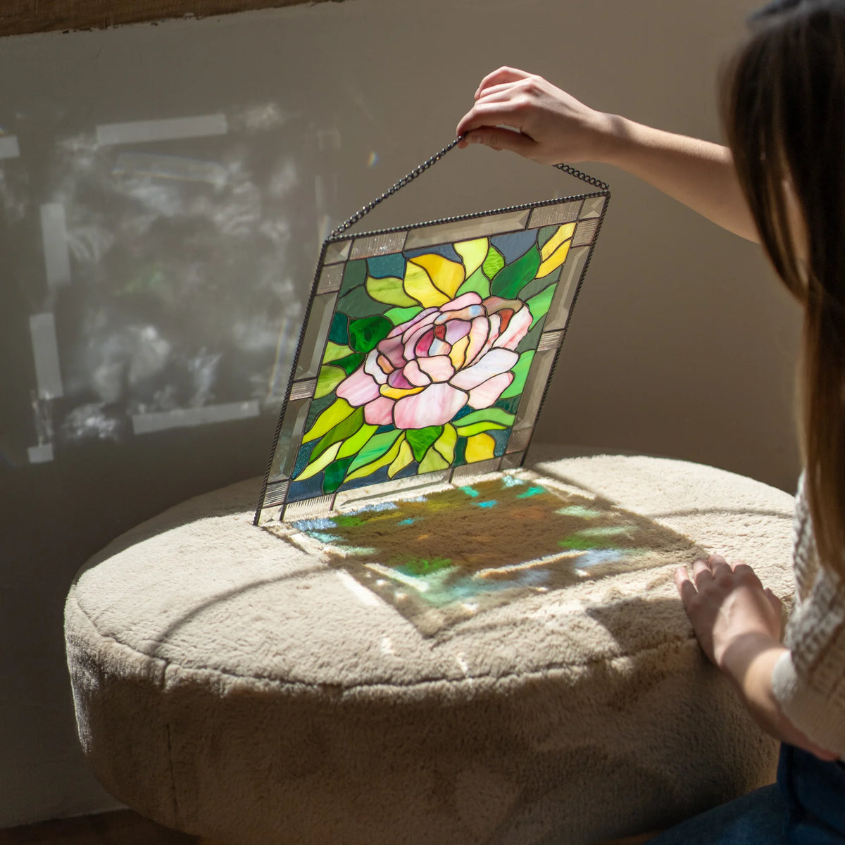Stained glass peony panel tilted on a cushioned ottoman, sunlight streaming through and casting colorful reflections on the fabric.
