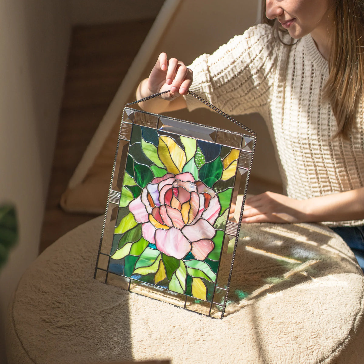 Woman smiling while holding a stained glass pink peony panel by its chain, sunlight illuminating the pink petals and green leaves.