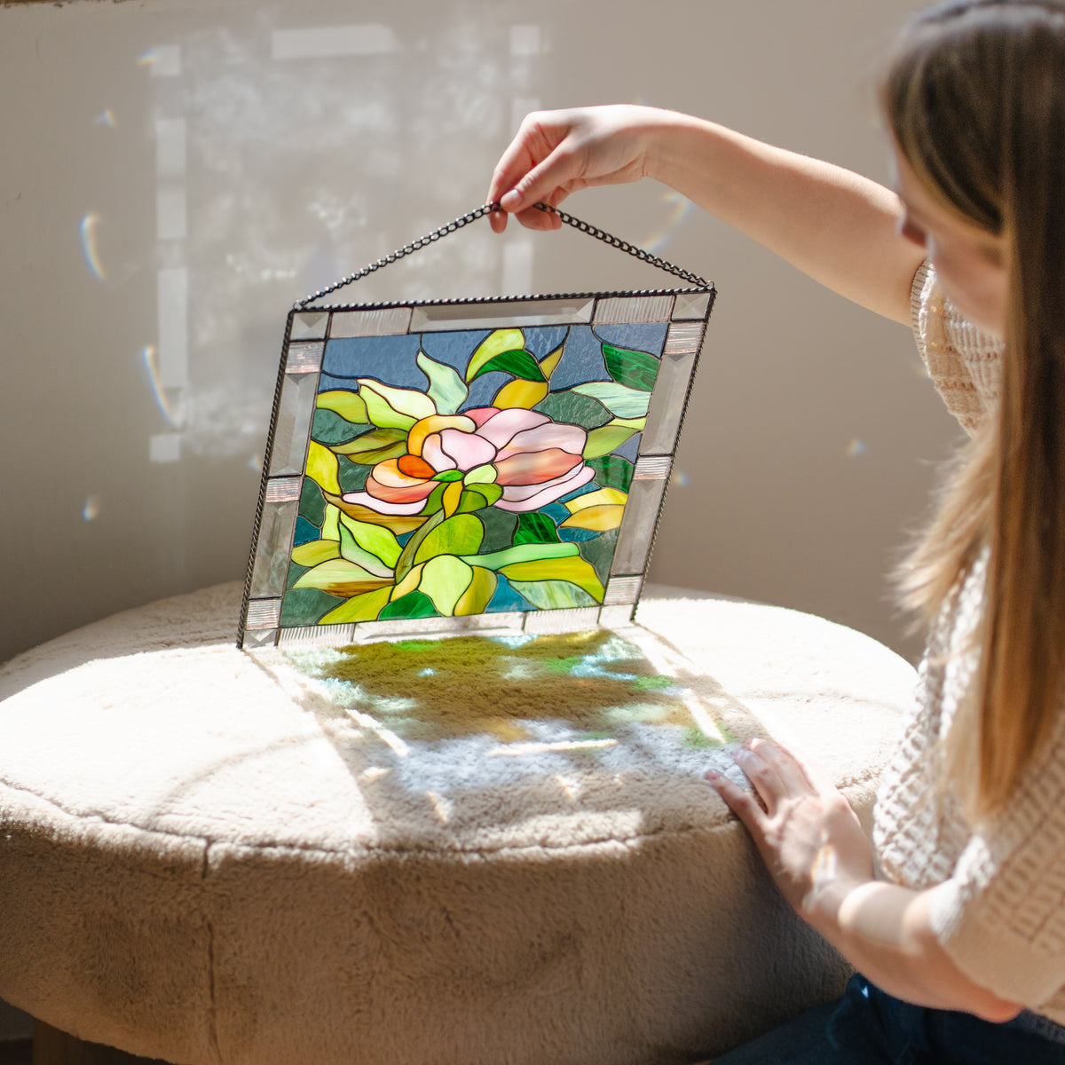 Stained glass peony flower panel with a beveled border hanging by a chain against a bright window, pink bloom and green leaves catching light.