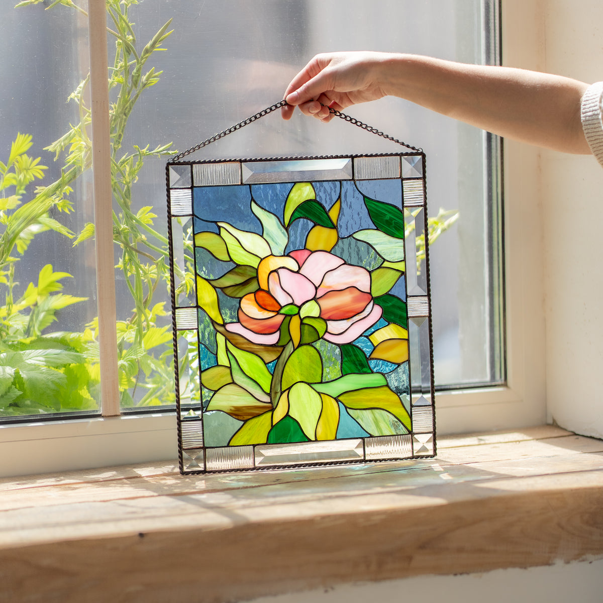 Hand lifting a stained glass peony panel by its chain in front of a sunlit window with green plants outside.