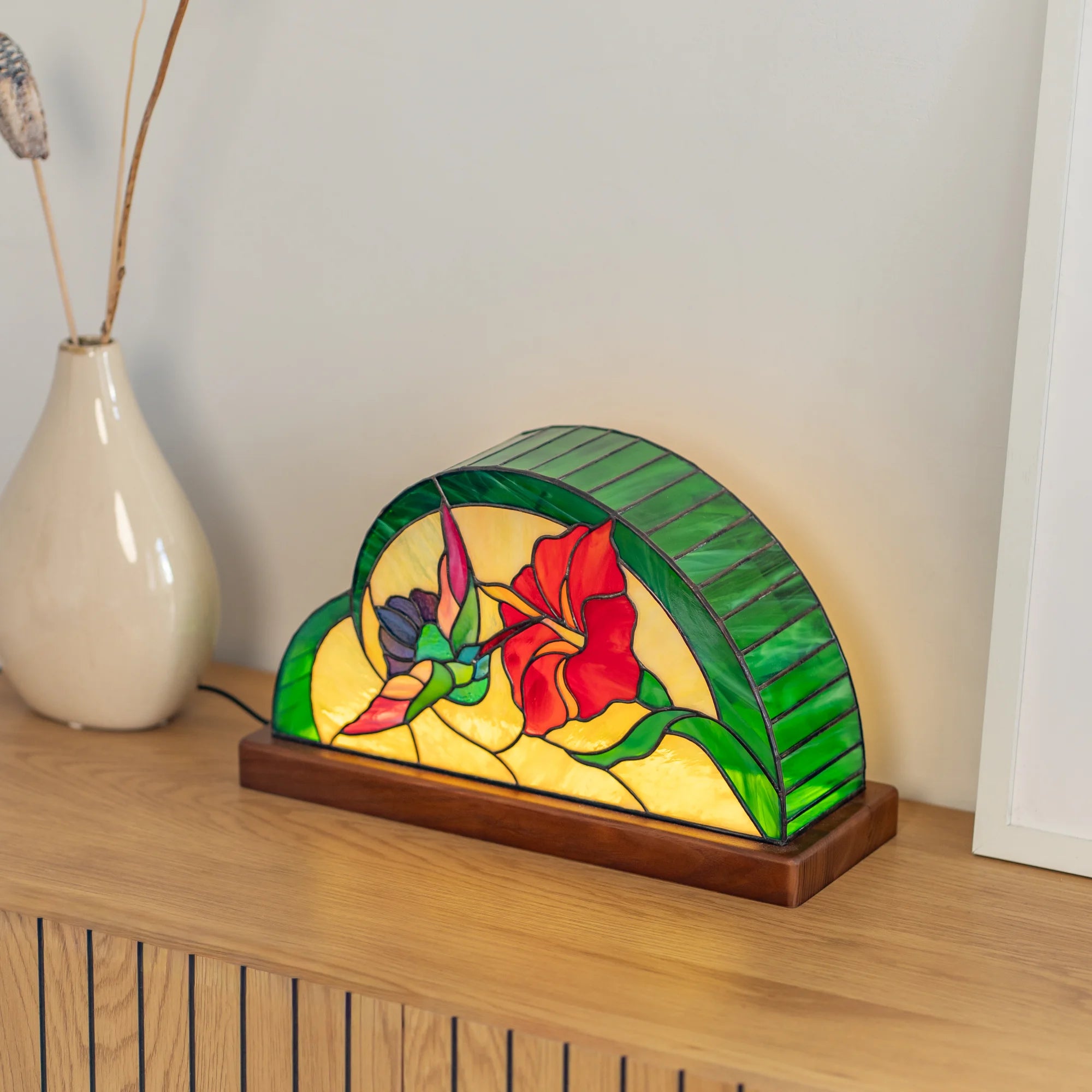 Hummingbird and hibiscus stained glass table lamp glowing on a wooden cabinet beside a cream vase - handmade Tiffany-technique lamp casting warm amber light in a cozy room