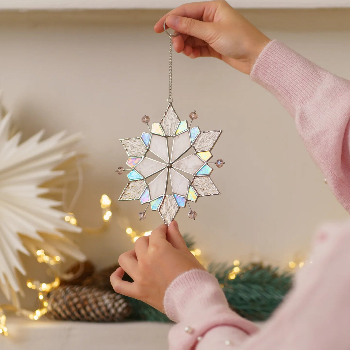 Woman in a pink sweater holding a snowflake with pink beads stained glass suncatcher in front of a paper star and fairy lights - handmade iridescent glass Christmas ornament and holiday gift