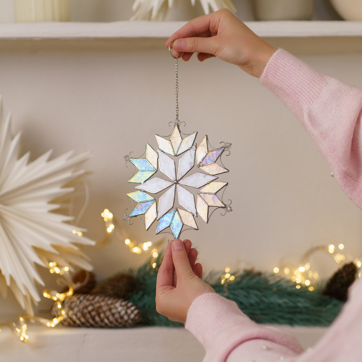 Woman in a pink sweater holding the iridescent snowflake with beads stained glass suncatcher in front of paper stars and fairy lights - handmade Christmas ornament and holiday gift