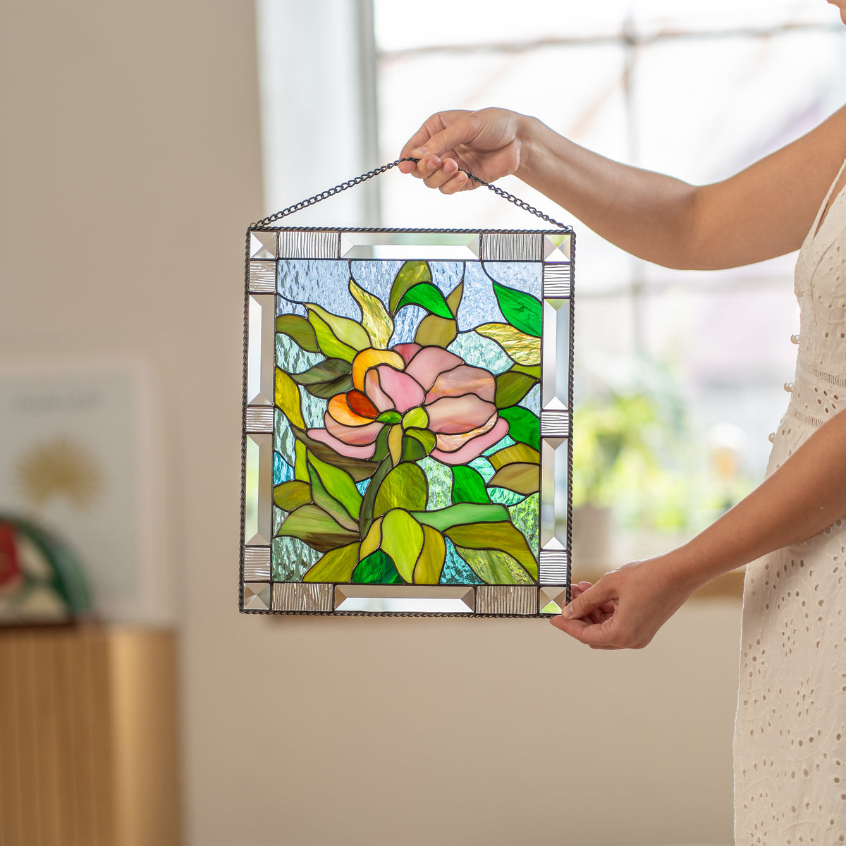 Woman looking down at a stained glass peony panel resting on a cushioned ottoman in a sunlit room.