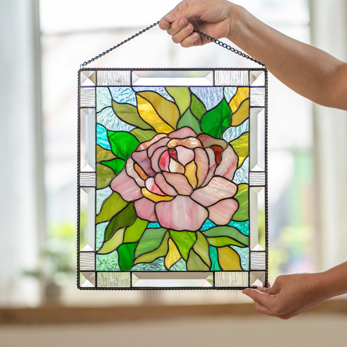 Stained glass pink peony panel held upright with both hands indoors near a staircase, light passing through the beveled glass border.