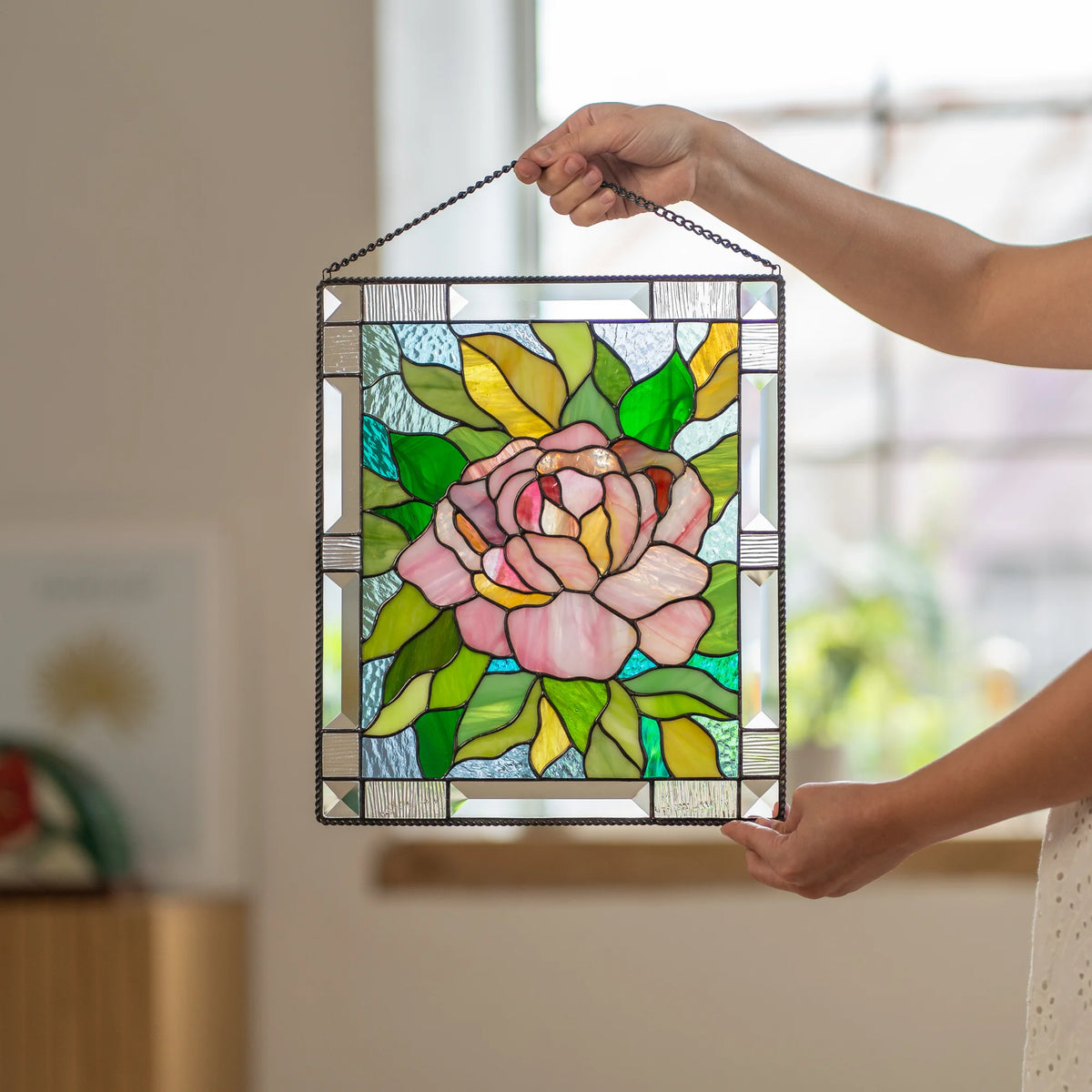 Woman standing and holding a stained glass pink peony panel with both hands, showing the full flower and leaf design up close.