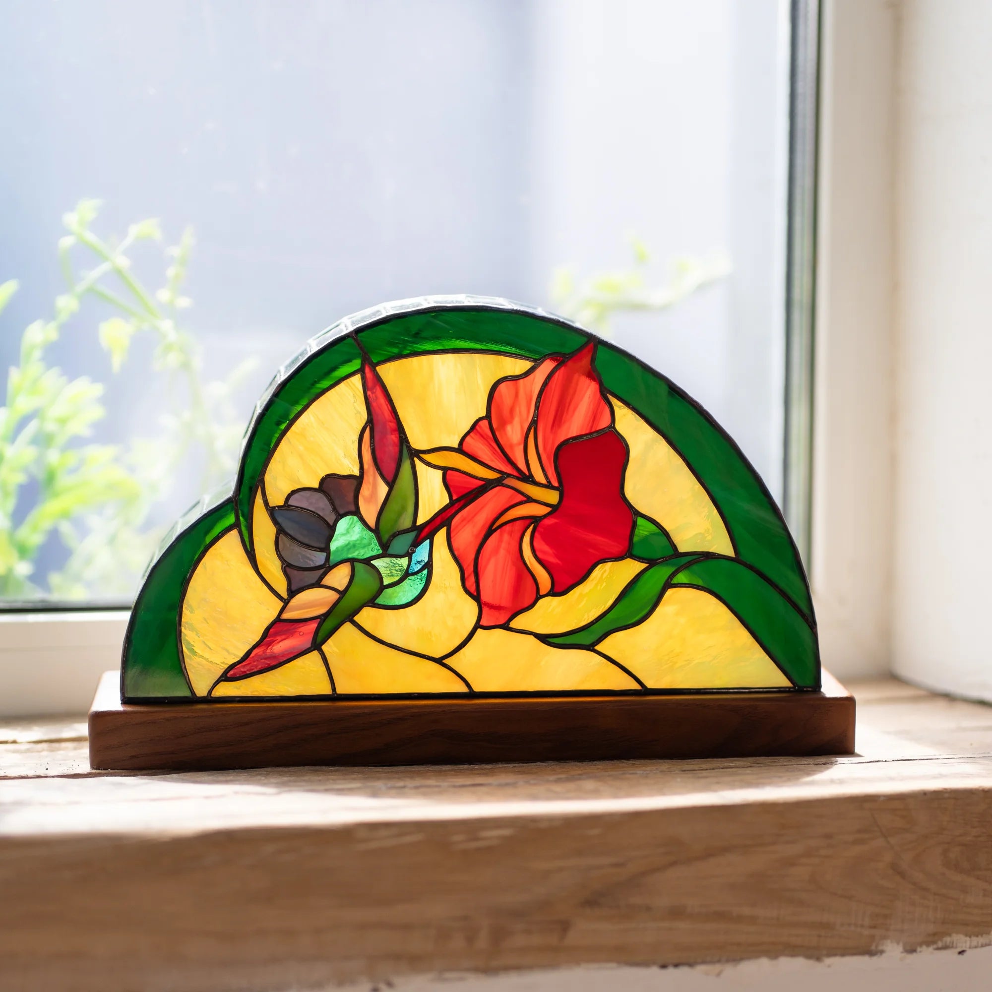 Hummingbird stained glass table lamp on a sunlit windowsill, a hummingbird sipping from a red hibiscus glowing in daylight - handmade Tiffany-technique lamp on a wooden base