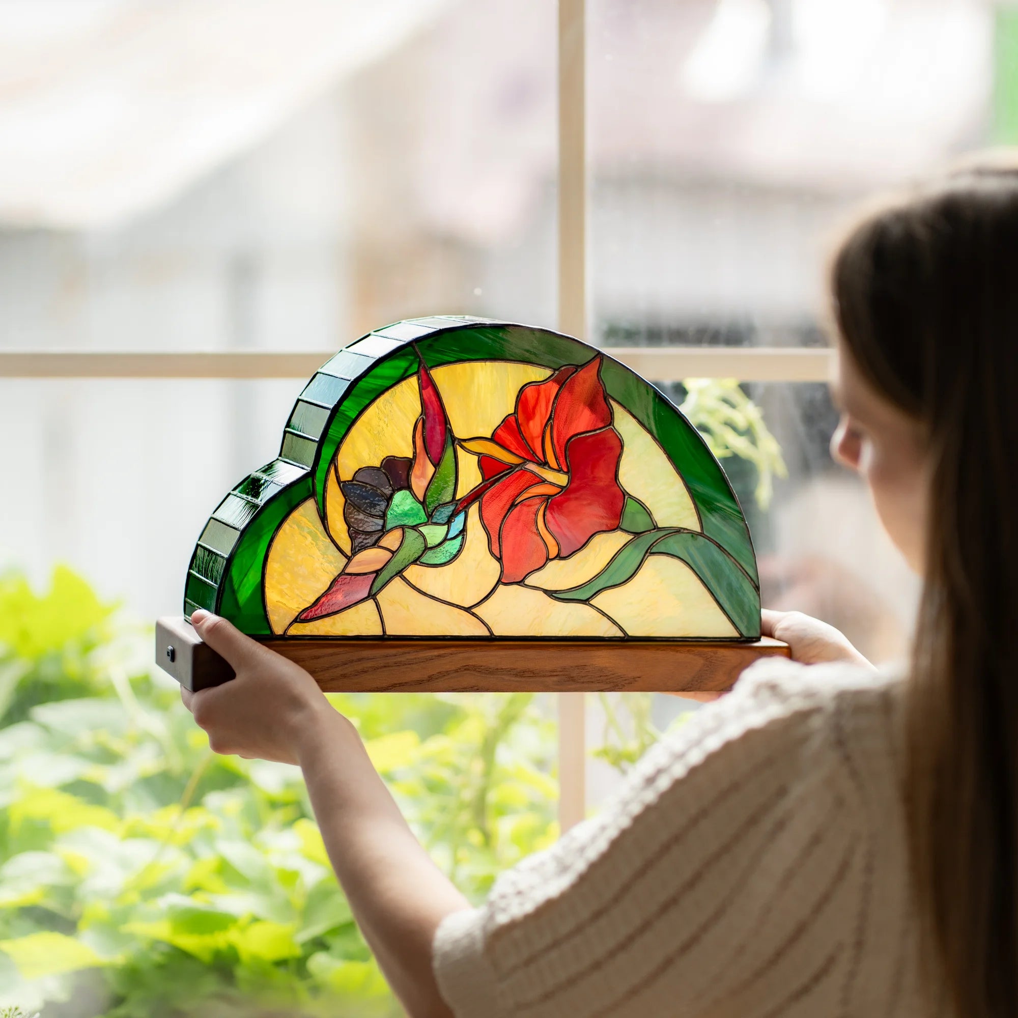 Woman holding a hummingbird stained glass table lamp up to a bright window, warm amber and green glass glowing against the light - handmade Tiffany-technique lamp and thoughtful gift
