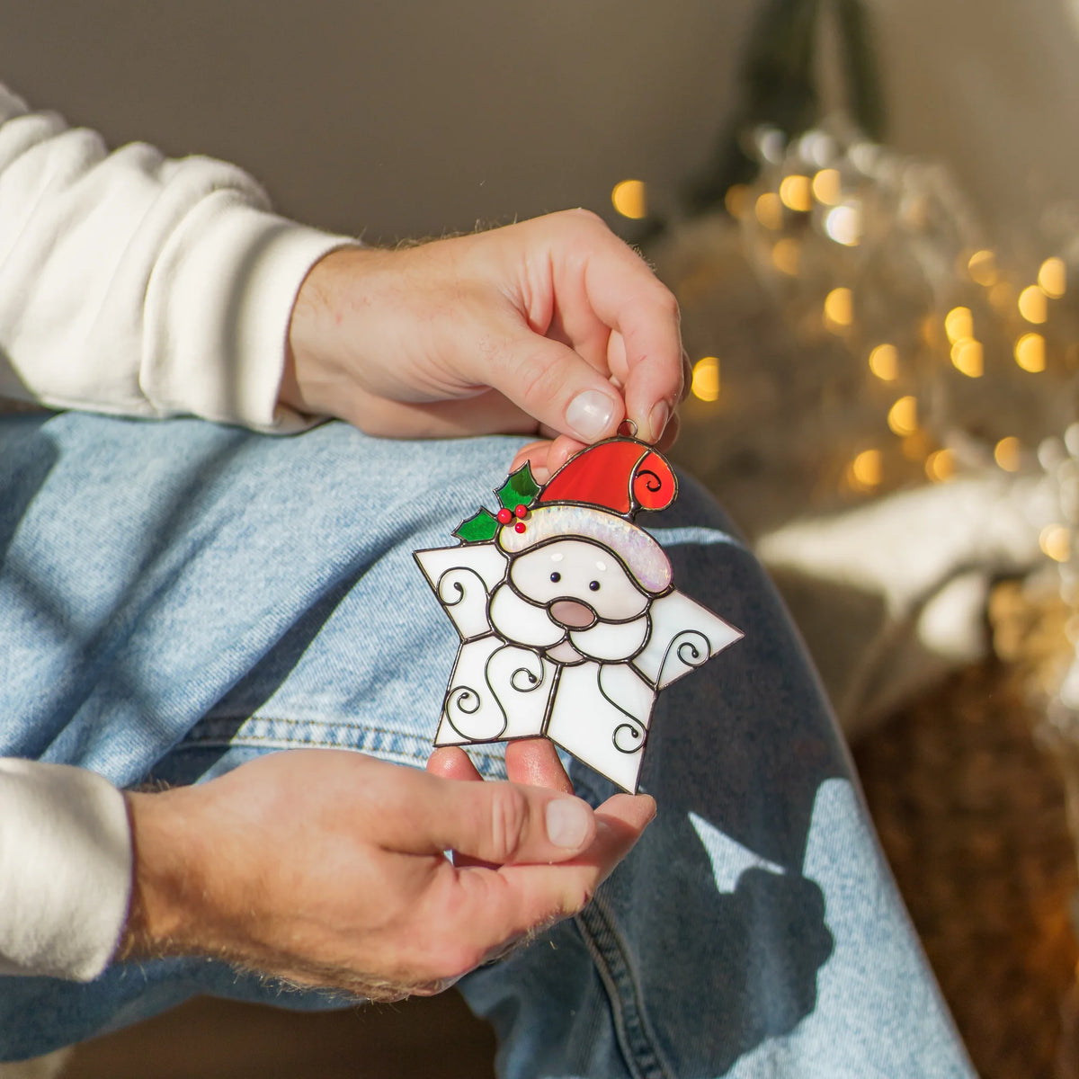 Hands holding the Snowflake Santa stained glass ornament by its loop above blue jeans, showing a white star beard with curly wire swirls and an iridescent hat brim - handmade Christmas decor