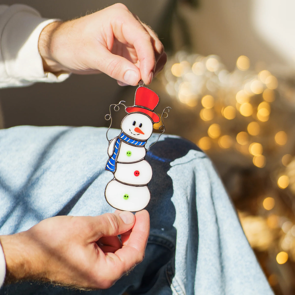 Hands holding the Christmas stained glass snowman ornament by its hanging loop, showing white glass body, red and green buttons and curly wire arms against warm bokeh lights - handmade Christmas decor