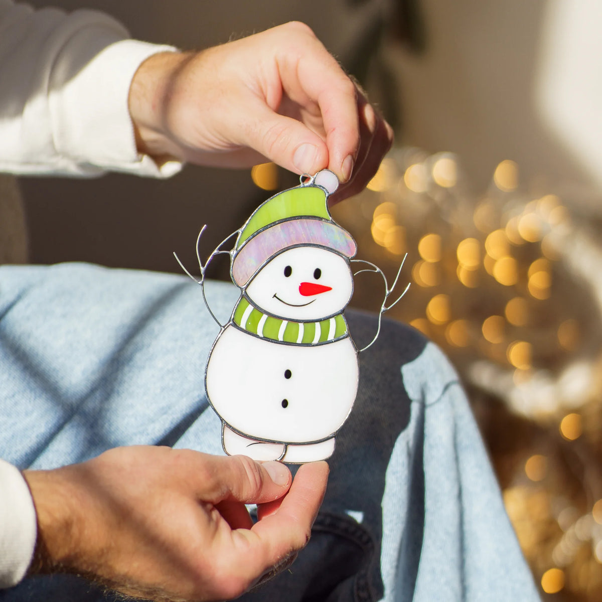 Man sitting on a couch with warm string lights in the background, holding a stained glass snowman suncatcher by its loop.