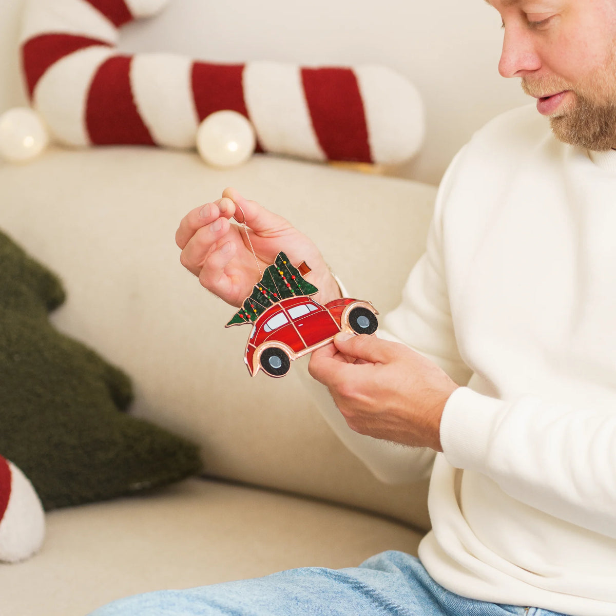 Man sitting on a couch surrounded by Christmas decor, holding and admiring a stained glass red car suncatcher with a tree on top.