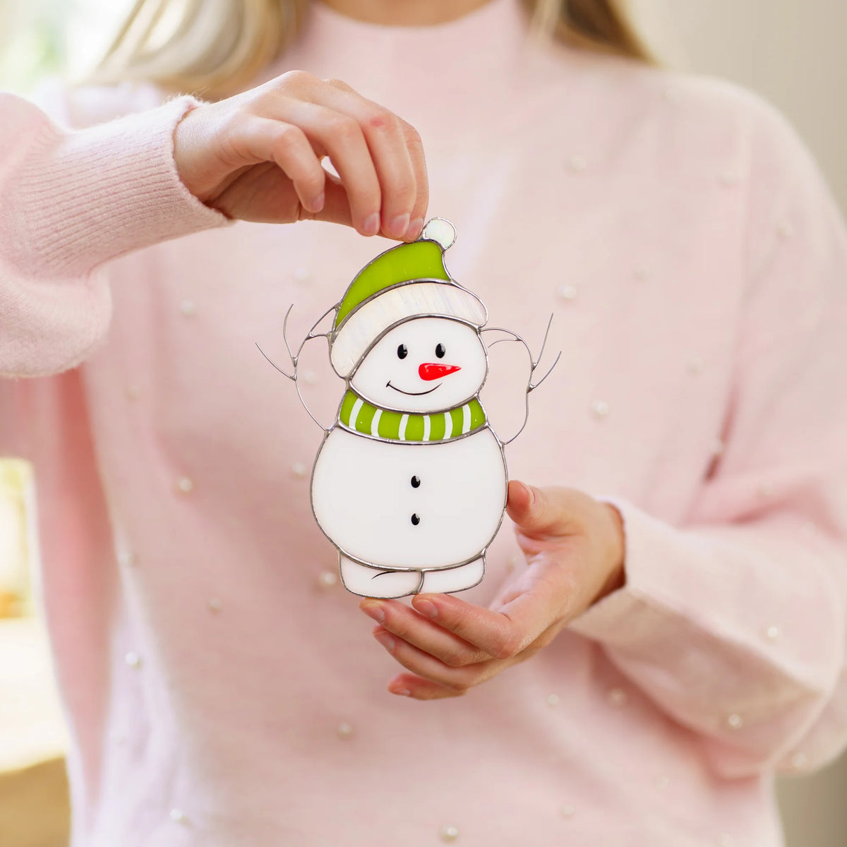 Woman in a pink sweater holding a stained glass snowman suncatcher with a green knit hat and striped scarf close in her hands.