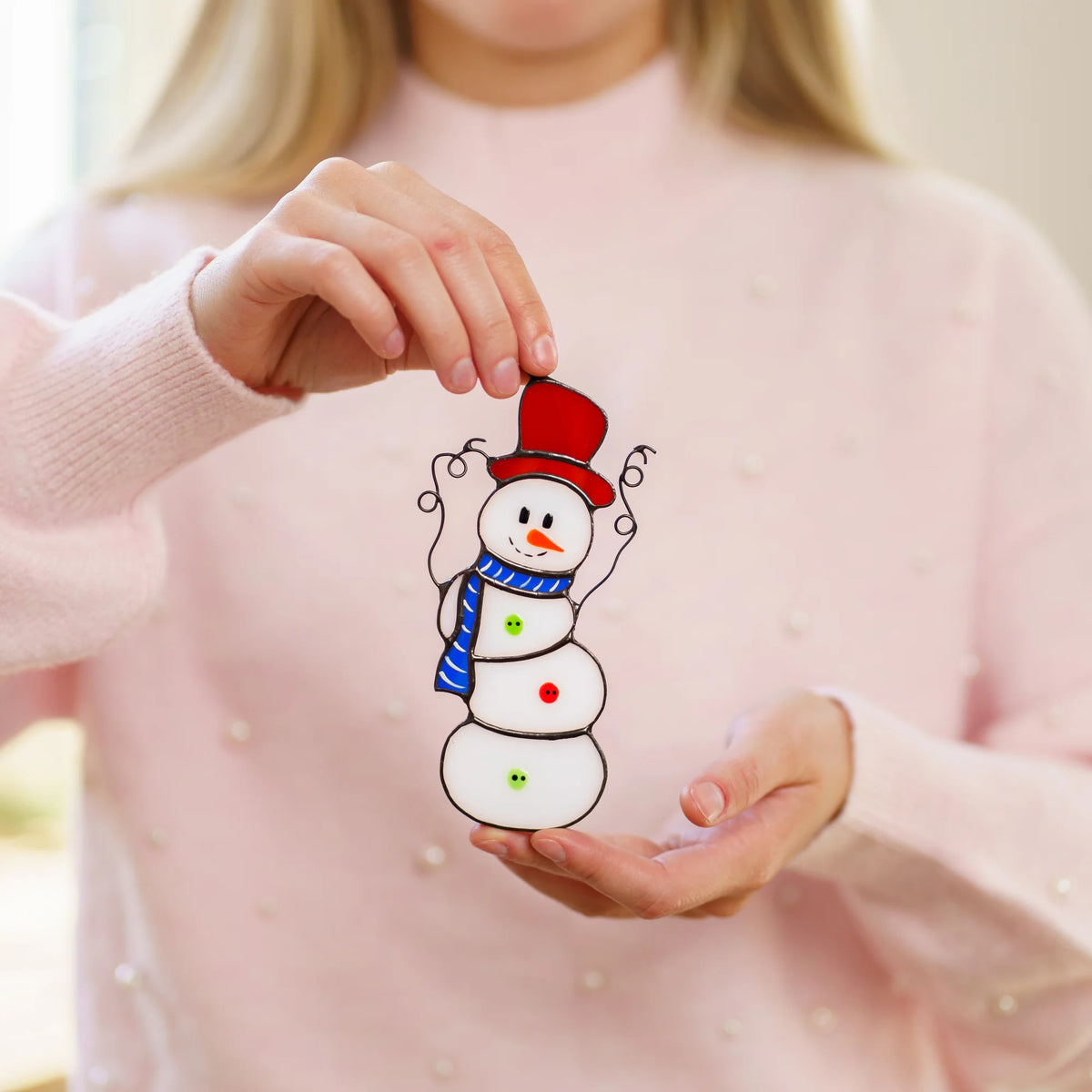 Woman in a pink sweater holding a handmade stained glass snowman with a red top hat, carrot nose and blue striped scarf - a cheerful Christmas suncatcher and a sweet holiday gift for her