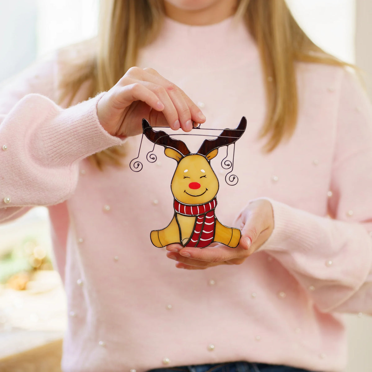 A person in a light pink sweater holds a small cartoon reindeer stained glass ornament with dark brown antlers and a red-and-white striped scarf.