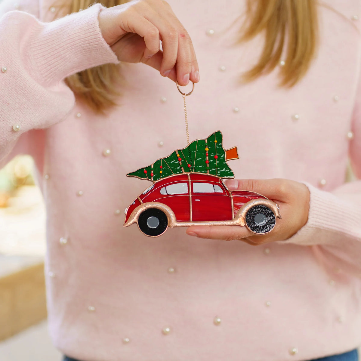 Woman in a pink sweater holding a stained glass suncatcher of a red car carrying a decorated Christmas tree in her hands.