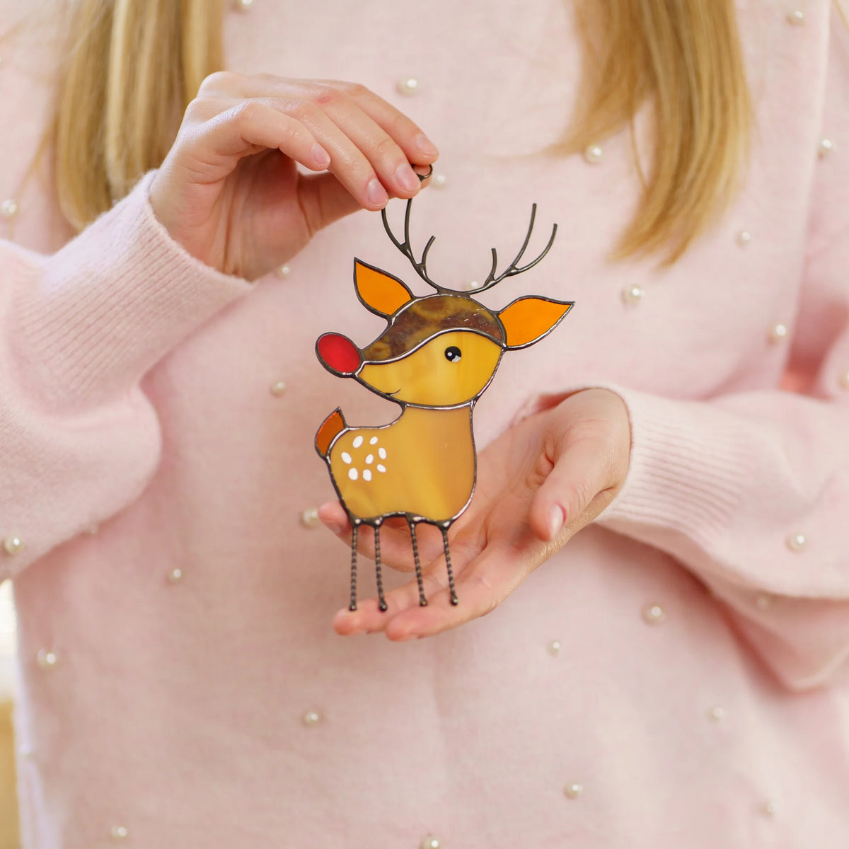 Woman in a pink pearl-studded sweater holding a stained glass reindeer suncatcher with a bright red nose close in her hands.