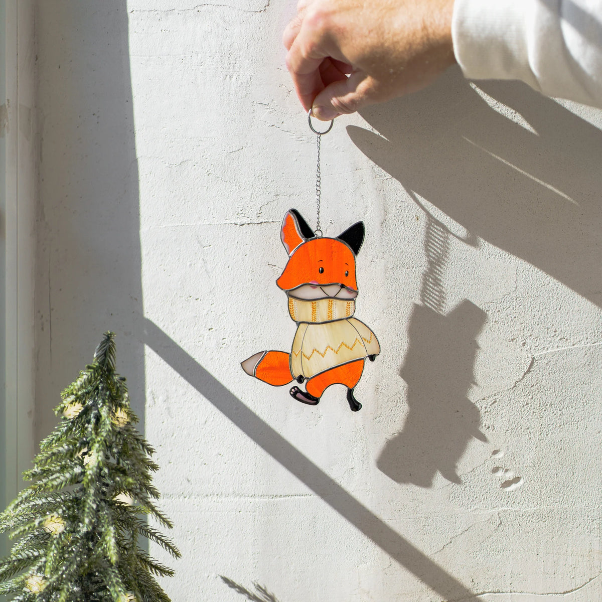 Close-up of two hands passing a stained glass fox suncatcher dressed in a cozy sweater, warm golden light catching the glass.