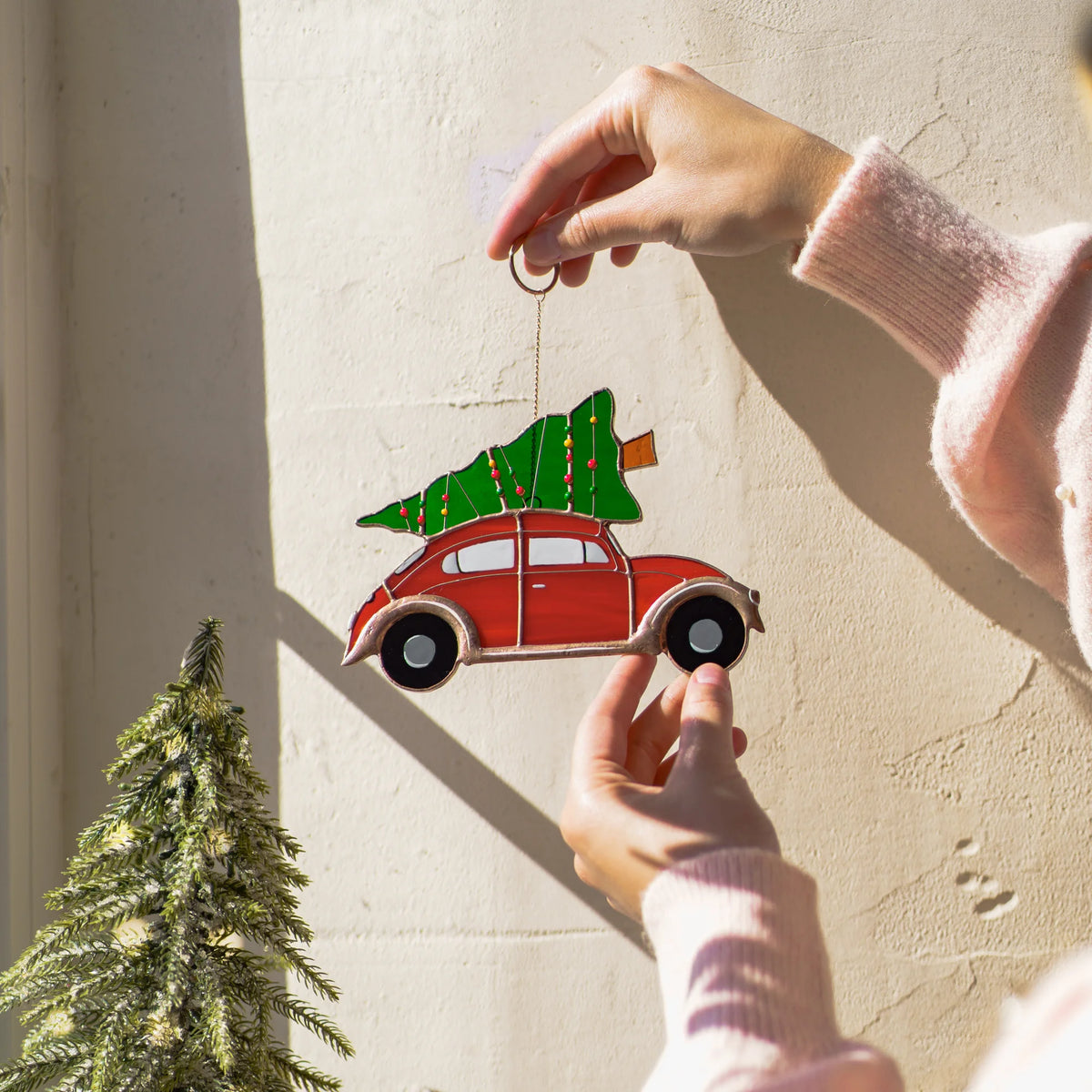 Hand holding the chain of a stained glass red car suncatcher against a sunlit window with a Christmas tree visible outside.