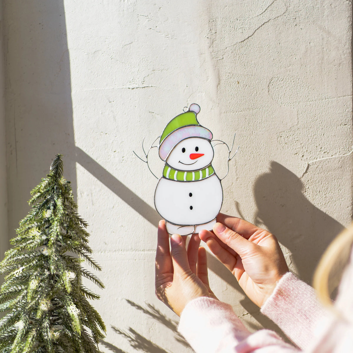 Hands holding a stained glass snowman suncatcher with a green hat and scarf outdoors against a sunlit stone wall and pine tree.