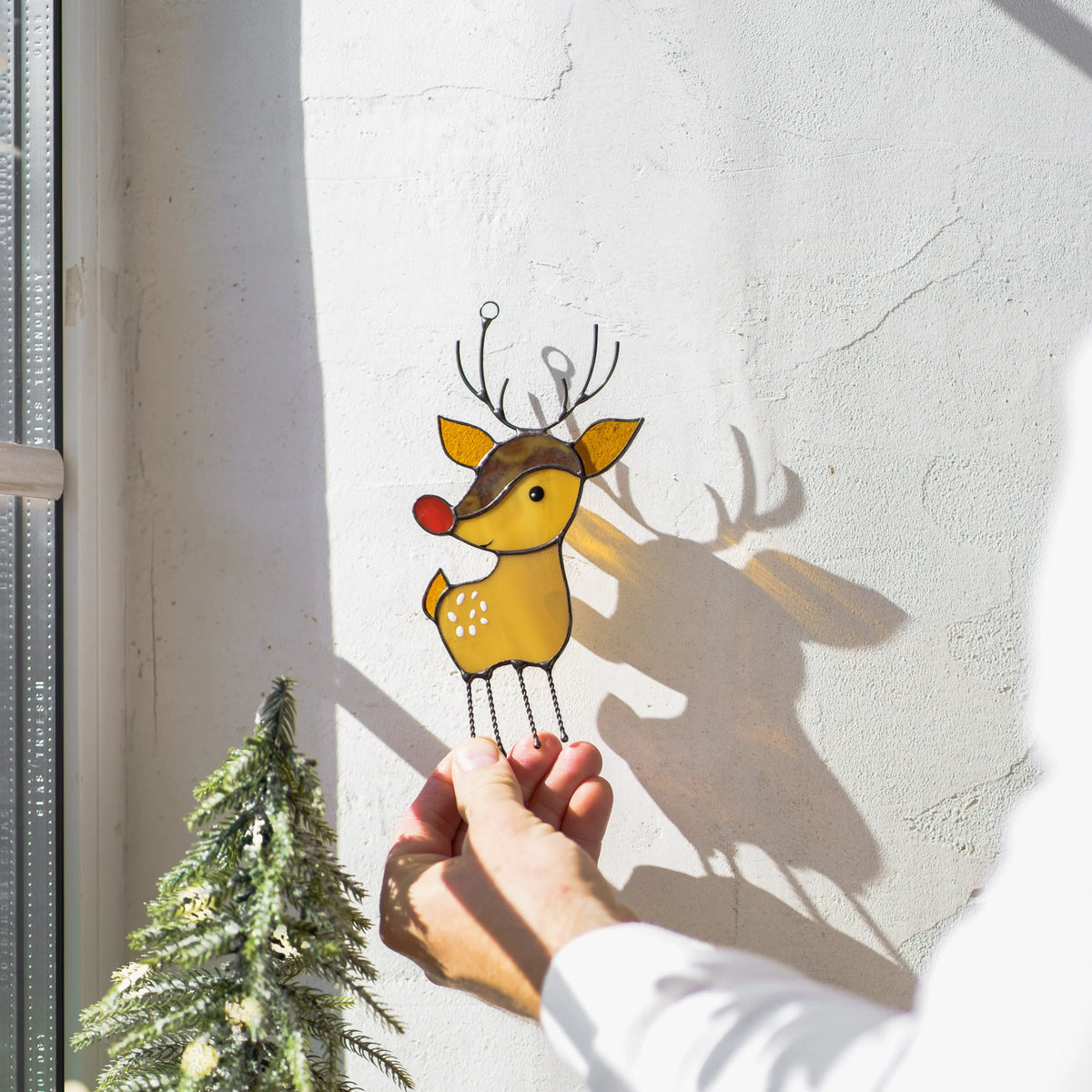 Hand holding the chain of a stained glass reindeer suncatcher against a sunlit white wall with a pine tree shadow nearby.