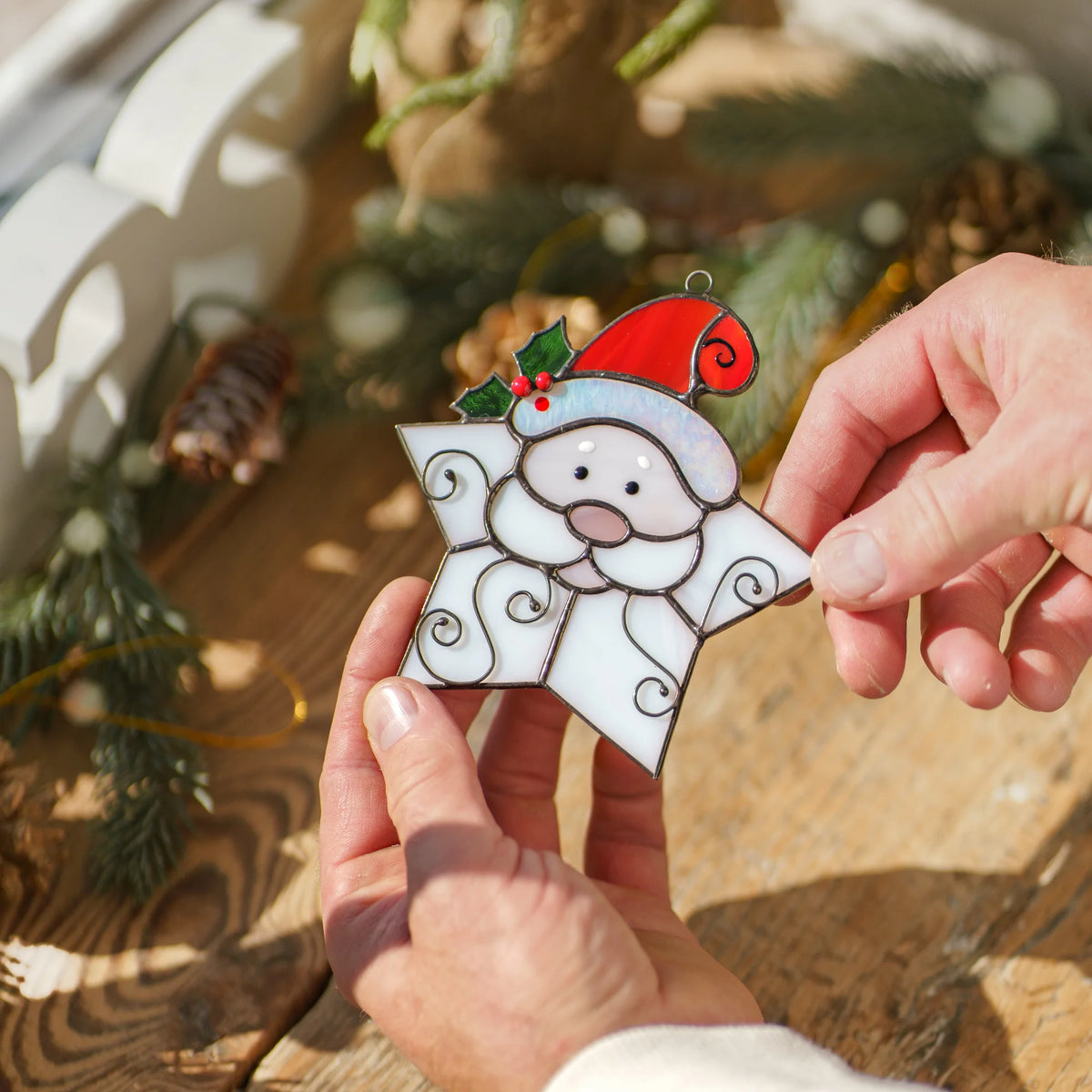Close-up of the Snowflake Santa stained glass ornament over a rustic wooden table with pine cones and fir branches, showing soldered seams, wire curls and painted holly berries - handmade Christmas suncatcher
