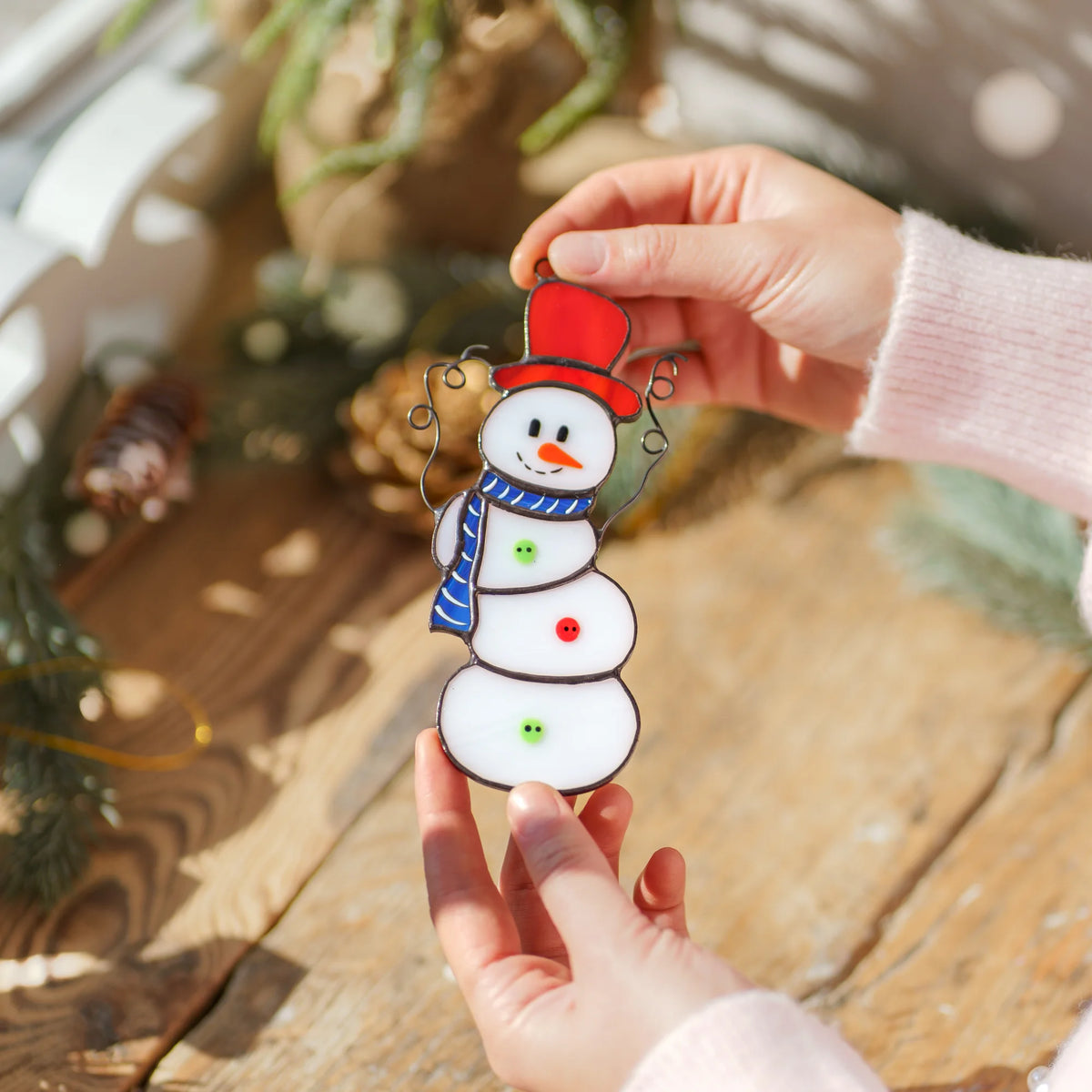 Close-up of the Christmas stained glass snowman over a rustic wooden table with pine cones and fir branches, showing red and green buttons and soldered seams - a handmade stained glass Christmas ornament